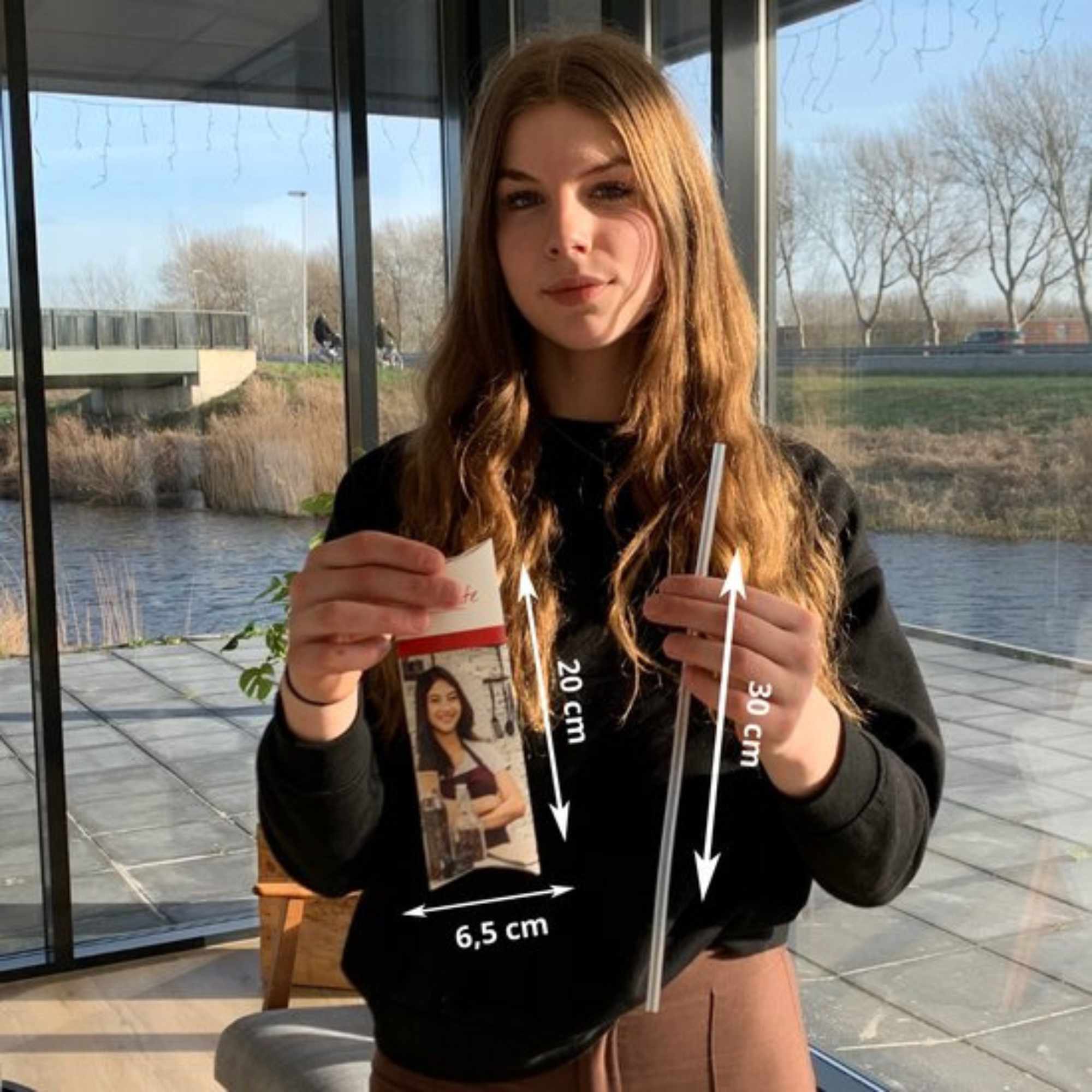 Woman holding a photo and a metal straw. The photo shows a woman. Background: a river, trees, and a bridge.