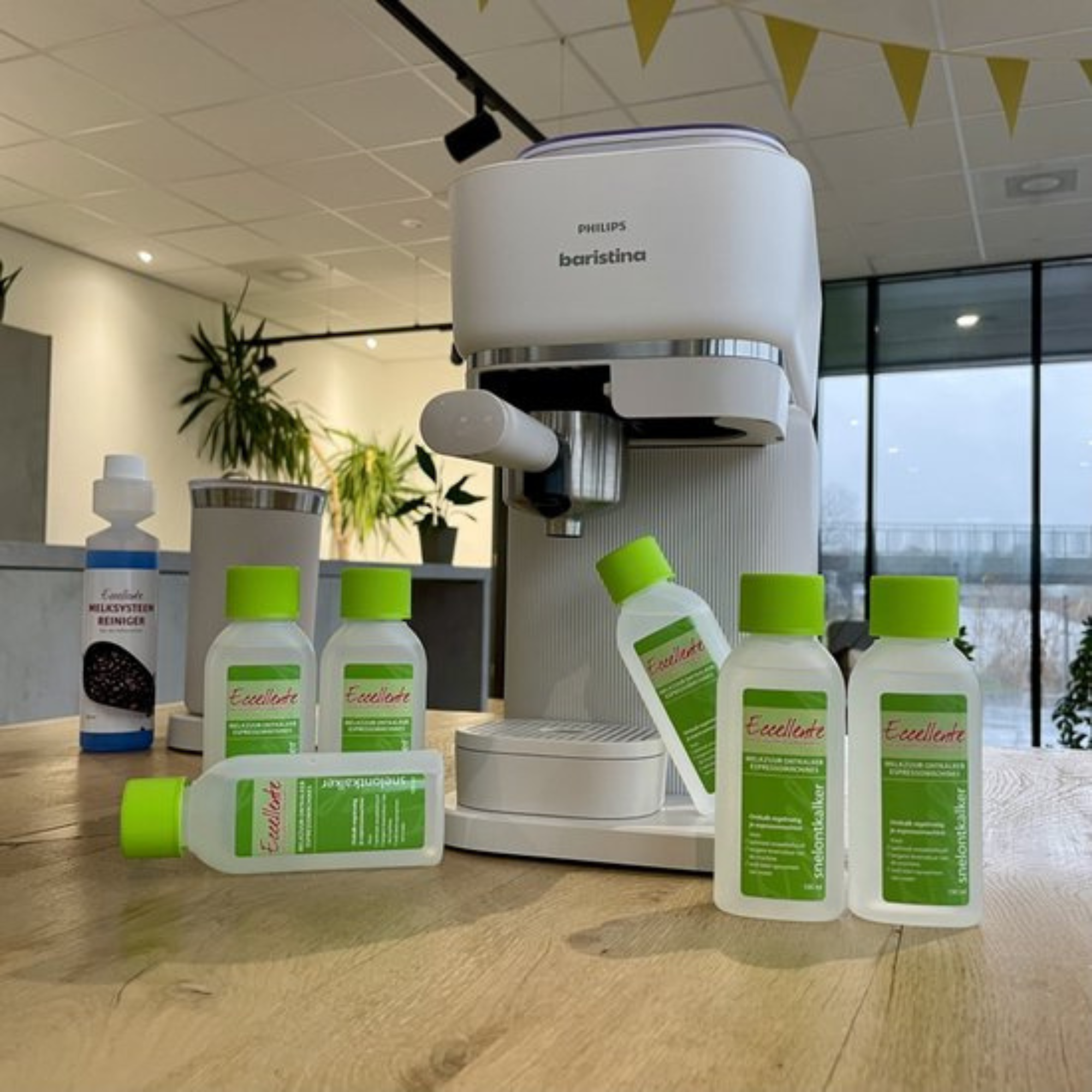 A white Philips Baristina espresso machine with cleaning products on a wooden table. Yellow flags hang above.
