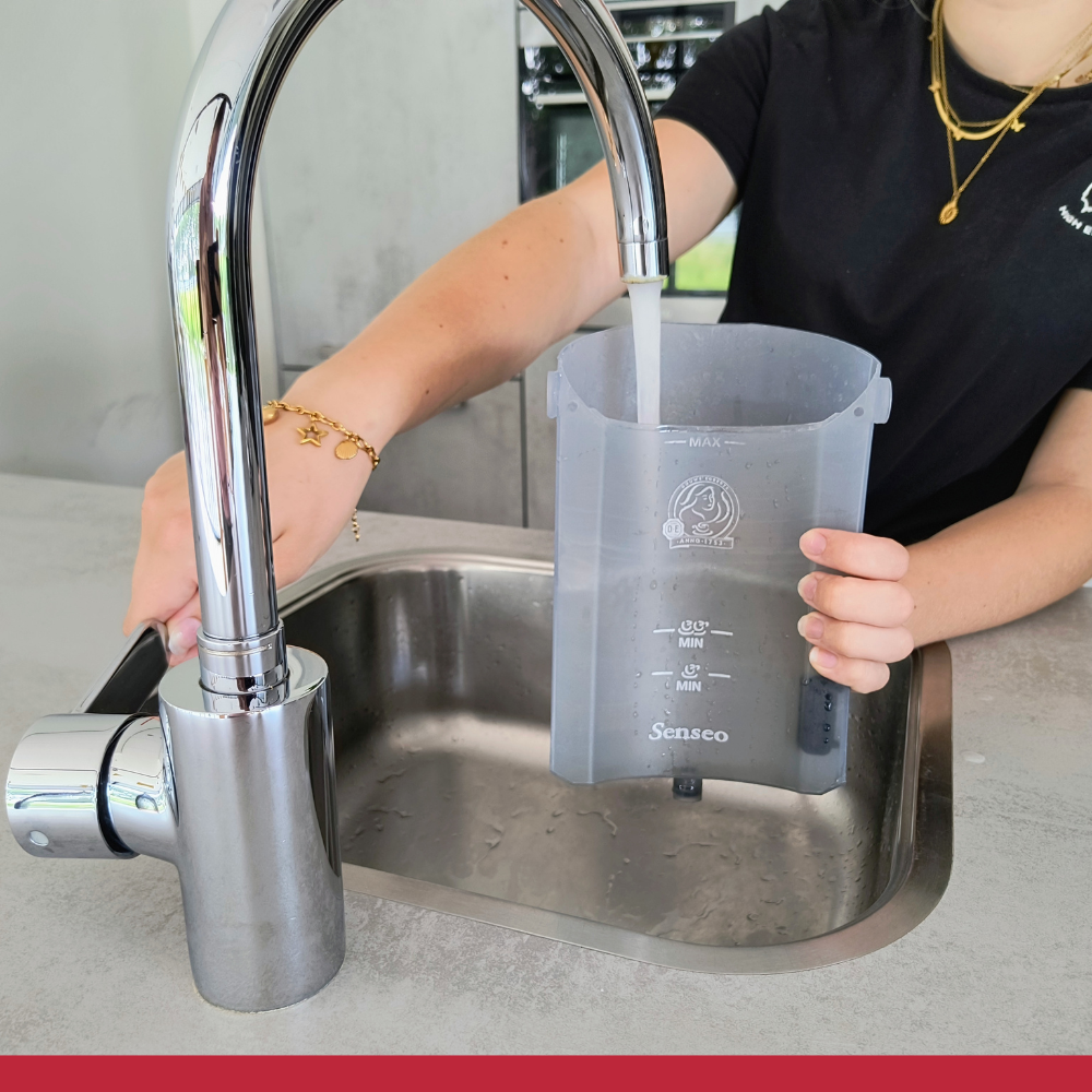 A person fills a transparent container with water from a chrome faucet. The container is in a stainless steel sink.
