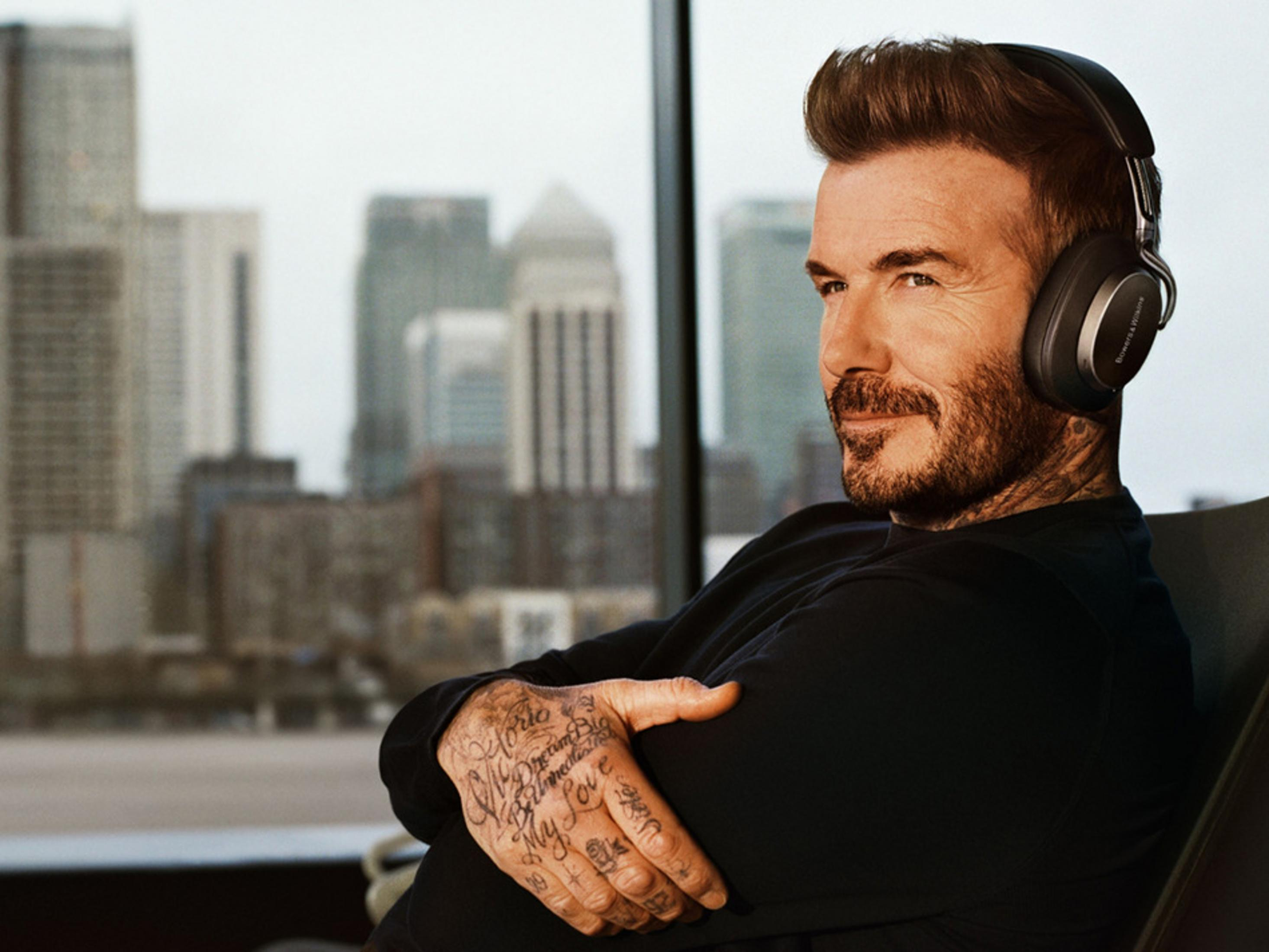 David Beckham w słuchawkach, patrzy w bok. Widoczne tatuaże na dłoniach i budynki miejskie w tle.
