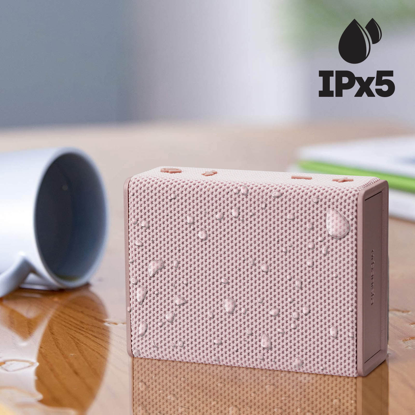 Różowy, wodoodporny głośnik z kroplami wody. Biały kubek po lewej. Widoczny symbol IPx5.