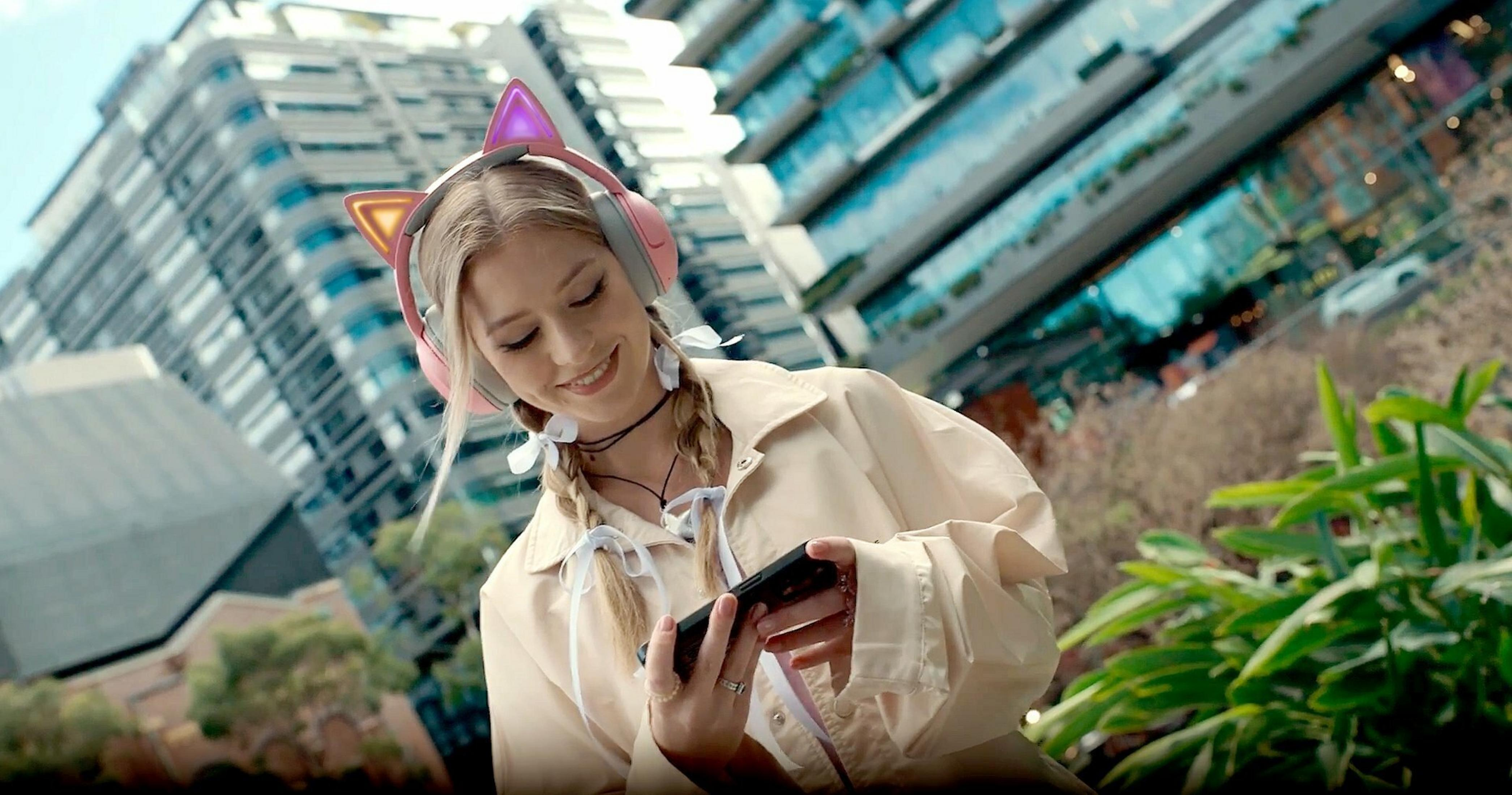 Kobieta w słuchawkach z kocimi uszami, trzymająca telefon, uśmiechnięta. Budynki i zieleń w tle.