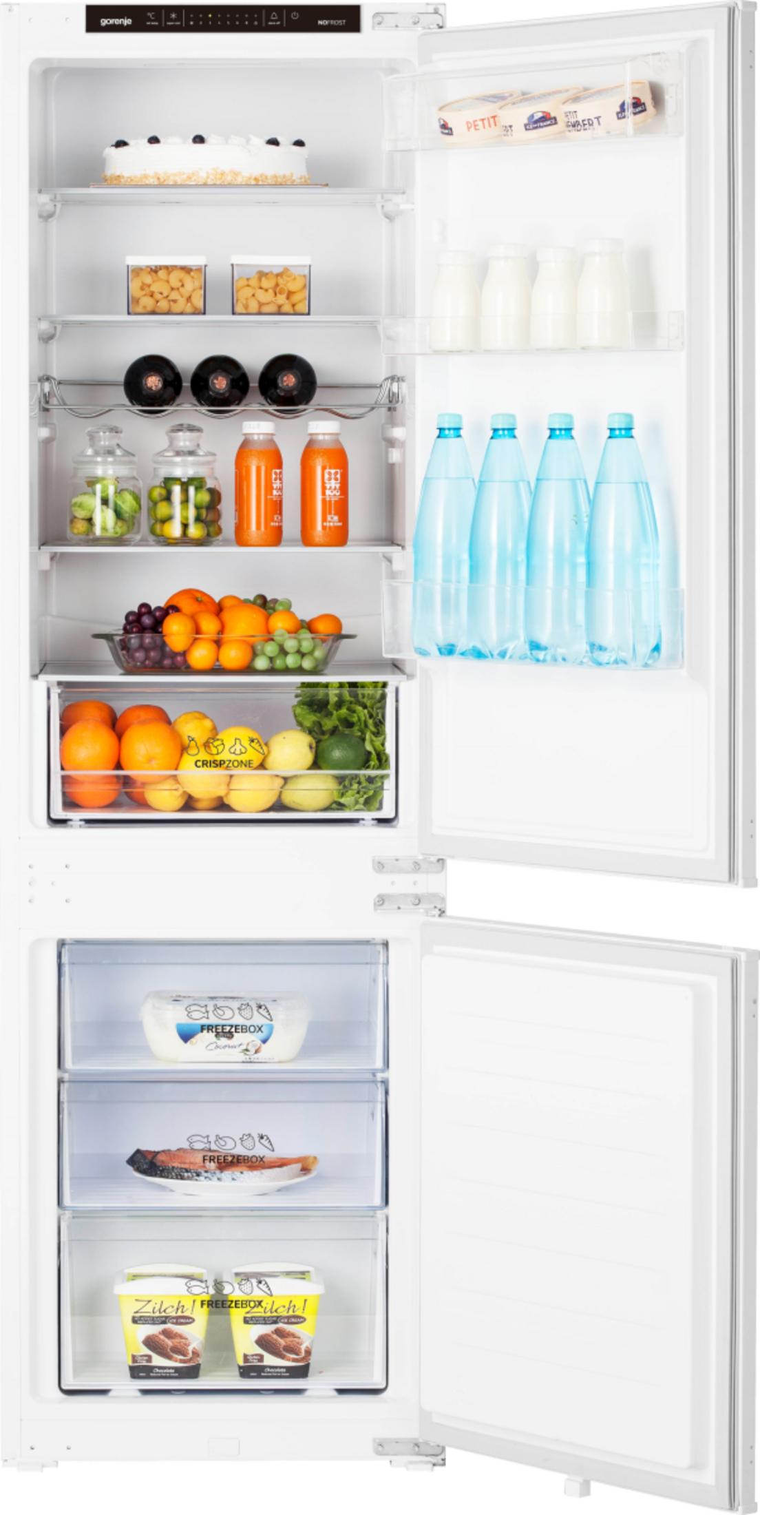 Otwarta lodówka wypełniona jedzeniem: ciasto, napoje, owoce, warzywa i mrożonki. Biała lodówka z otwartymi drzwiami.