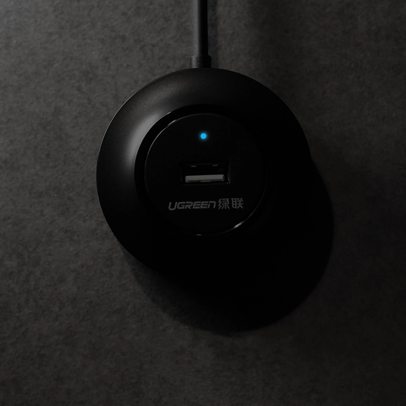 Czarny hub USB UGREEN z niebieskim światłem jest wyśrodkowany na ciemnoszarym, teksturowanym tle.