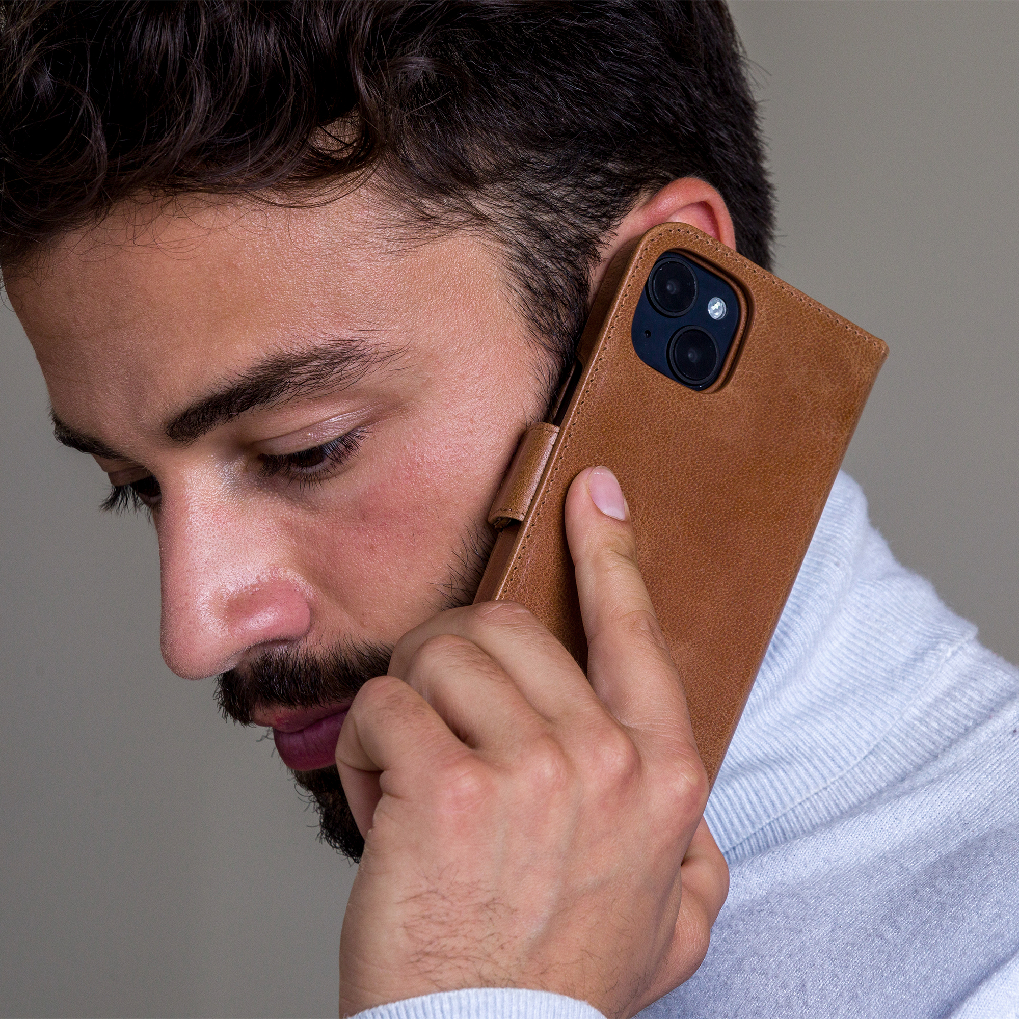 Un uomo con la barba tiene un telefono marrone all'orecchio. Indossa un maglione azzurro chiaro su sfondo grigio.
