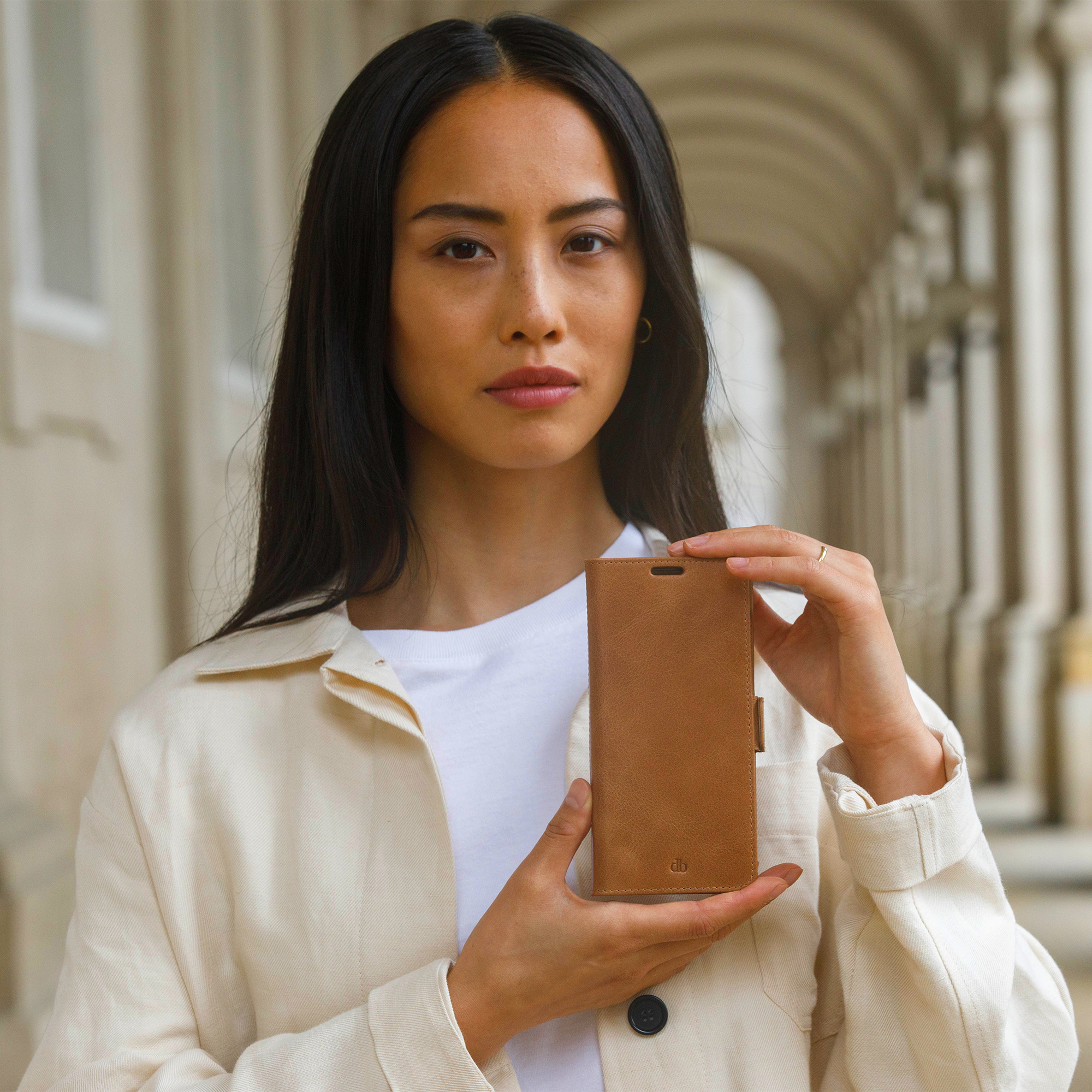 Una donna tiene in mano una custodia marrone per telefono. Indossa una giacca color crema e una camicia bianca. Lo sfondo è un edificio bianco.