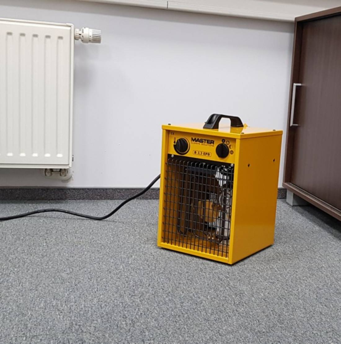 Een gele elektrische kachel staat op een grijs tapijt. Links een witte radiator. Rechts een bruine deur.