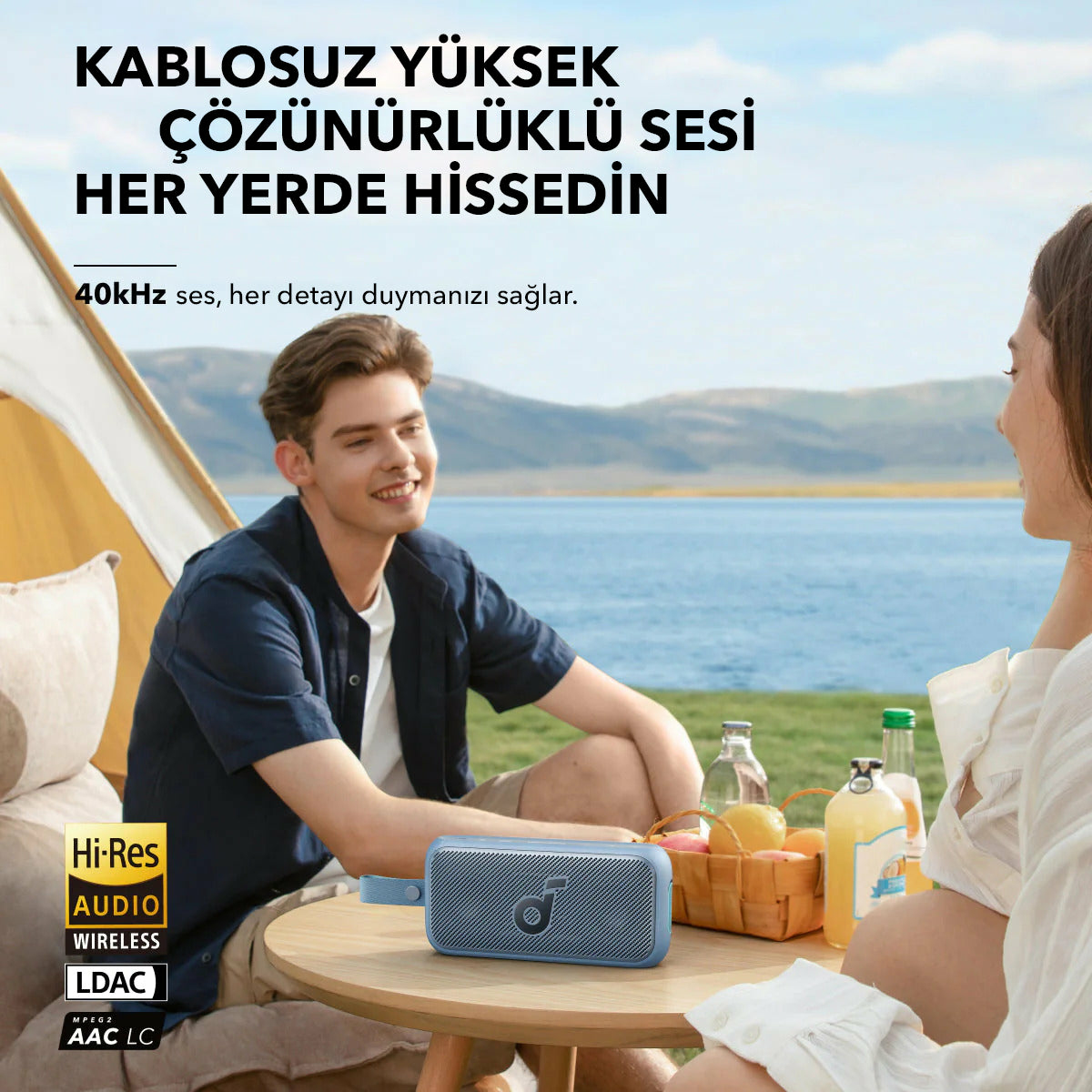 Mężczyzna i kobieta siedzą nad jeziorem z głośnikiem. Na stole owoce i napoje. Tekst: Bezprzewodowy dźwięk wysokiej rozdzielczości.