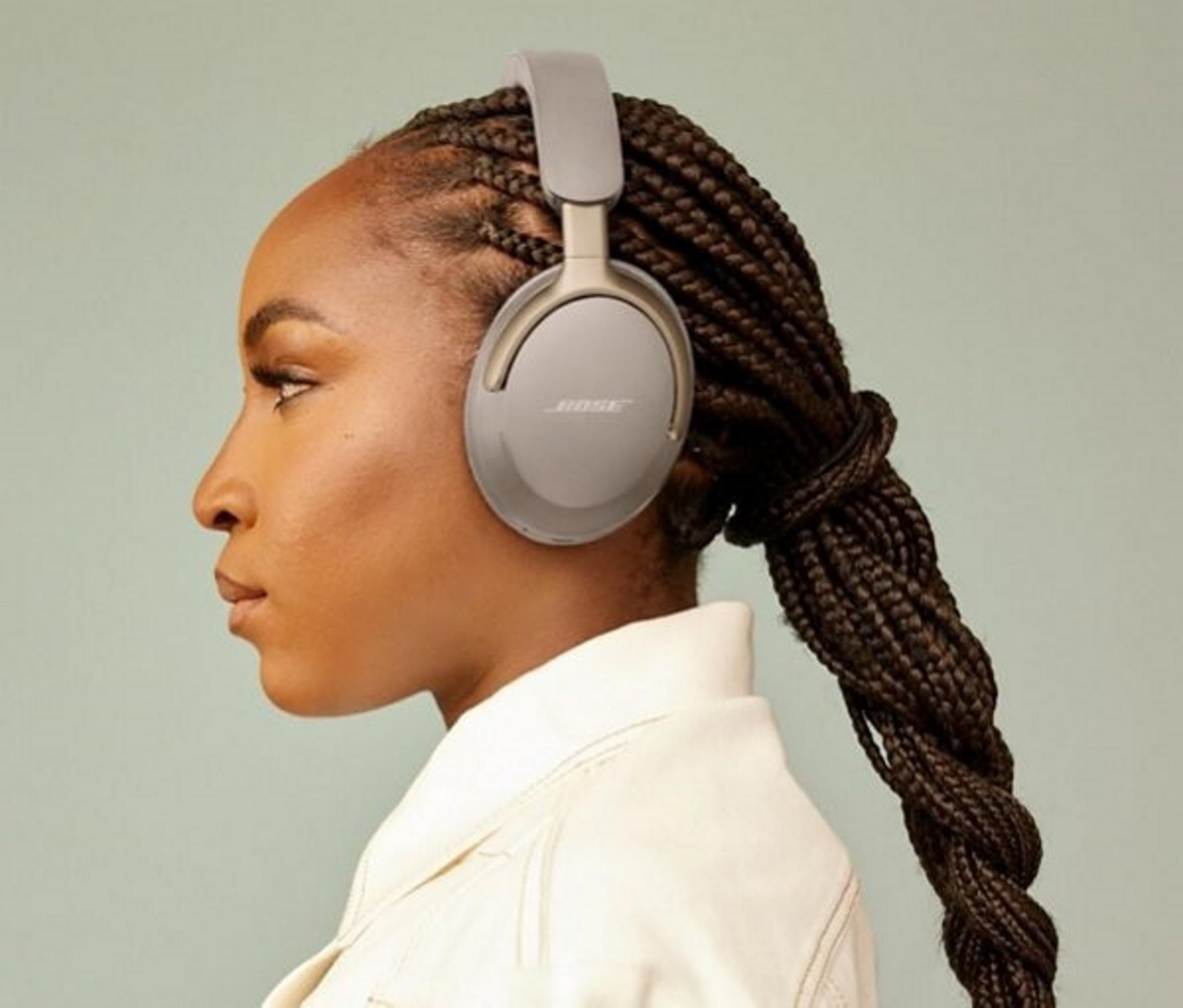 Kobieta z warkoczami w szarych słuchawkach Bose. Ma na sobie białą kurtkę. Tło jest jasnozielone.