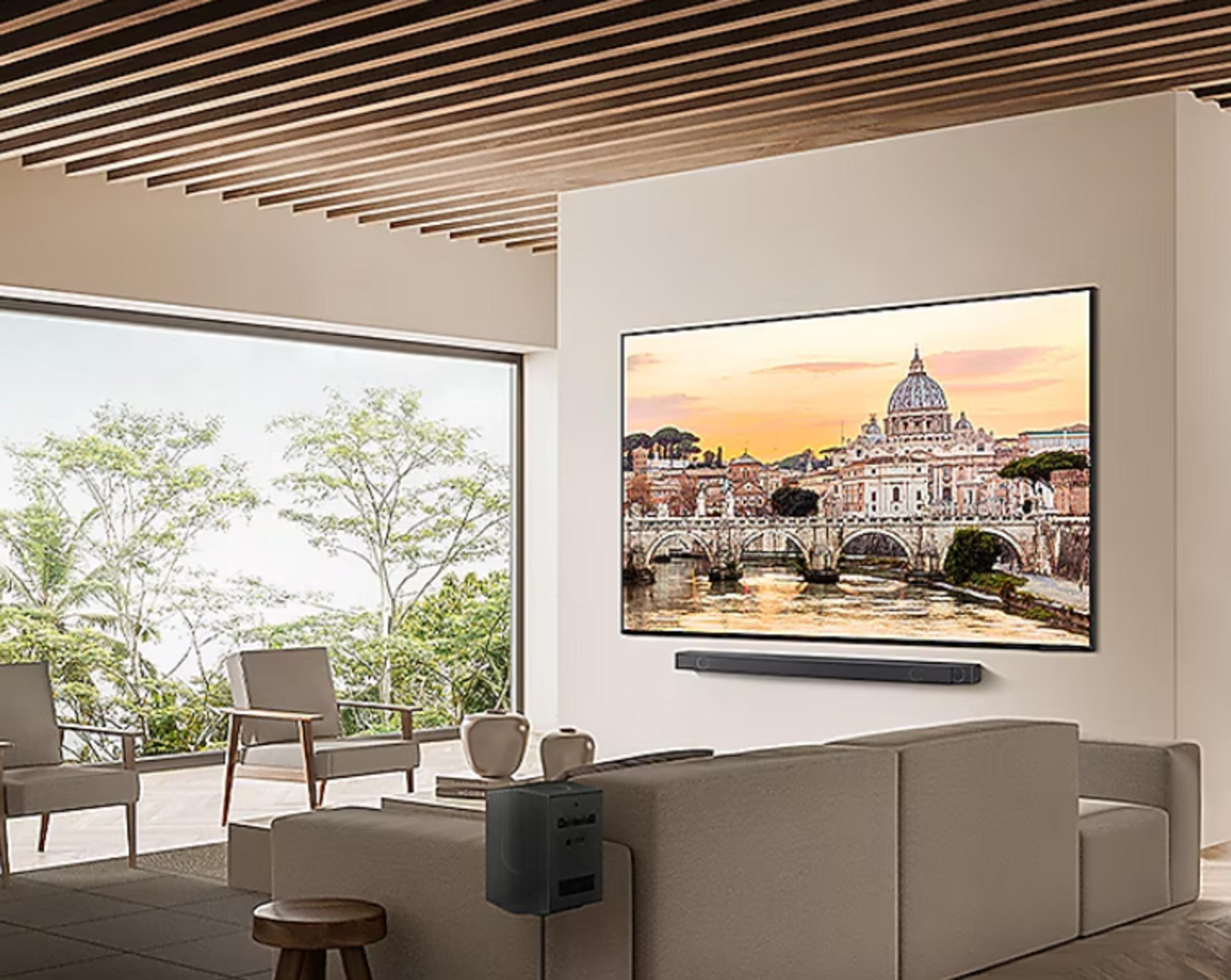 Nowoczesny salon z dużym telewizorem wyświetlającym krajobraz miejski. Widoczne są sofy, krzesła i duże okno z drzewami.