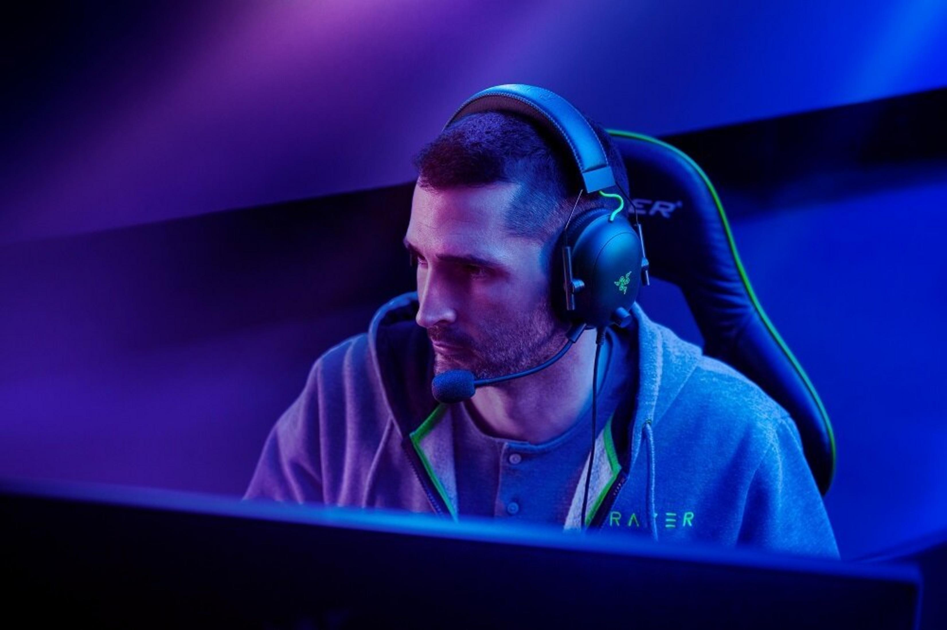 Hombre con auriculares, frente a una computadora, en una silla gamer. La escena está iluminada en azul y púrpura.