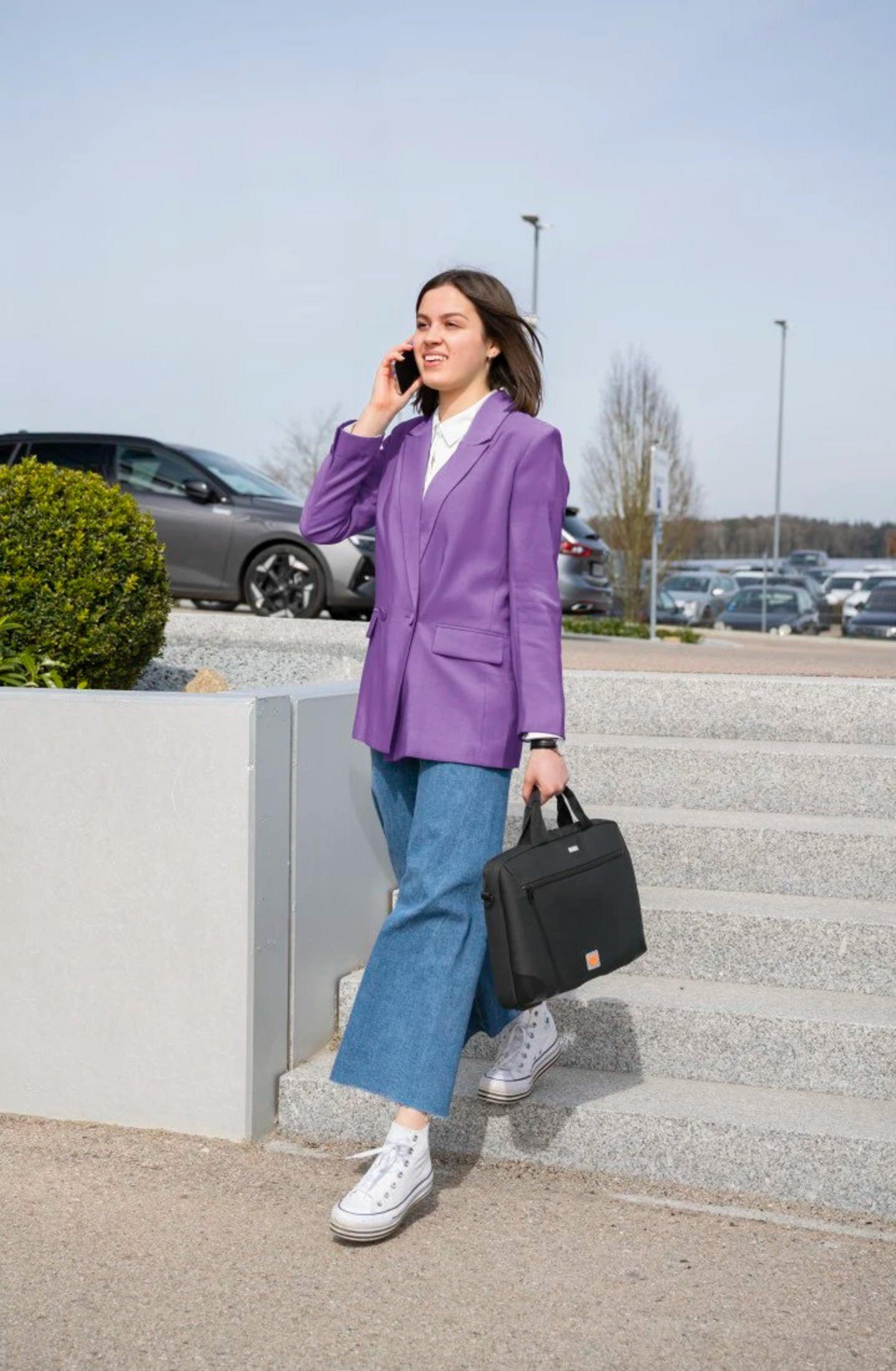 Una donna con un blazer viola e jeans scende le scale parlando al telefono e tenendo una borsa. Auto e alberi sullo sfondo.