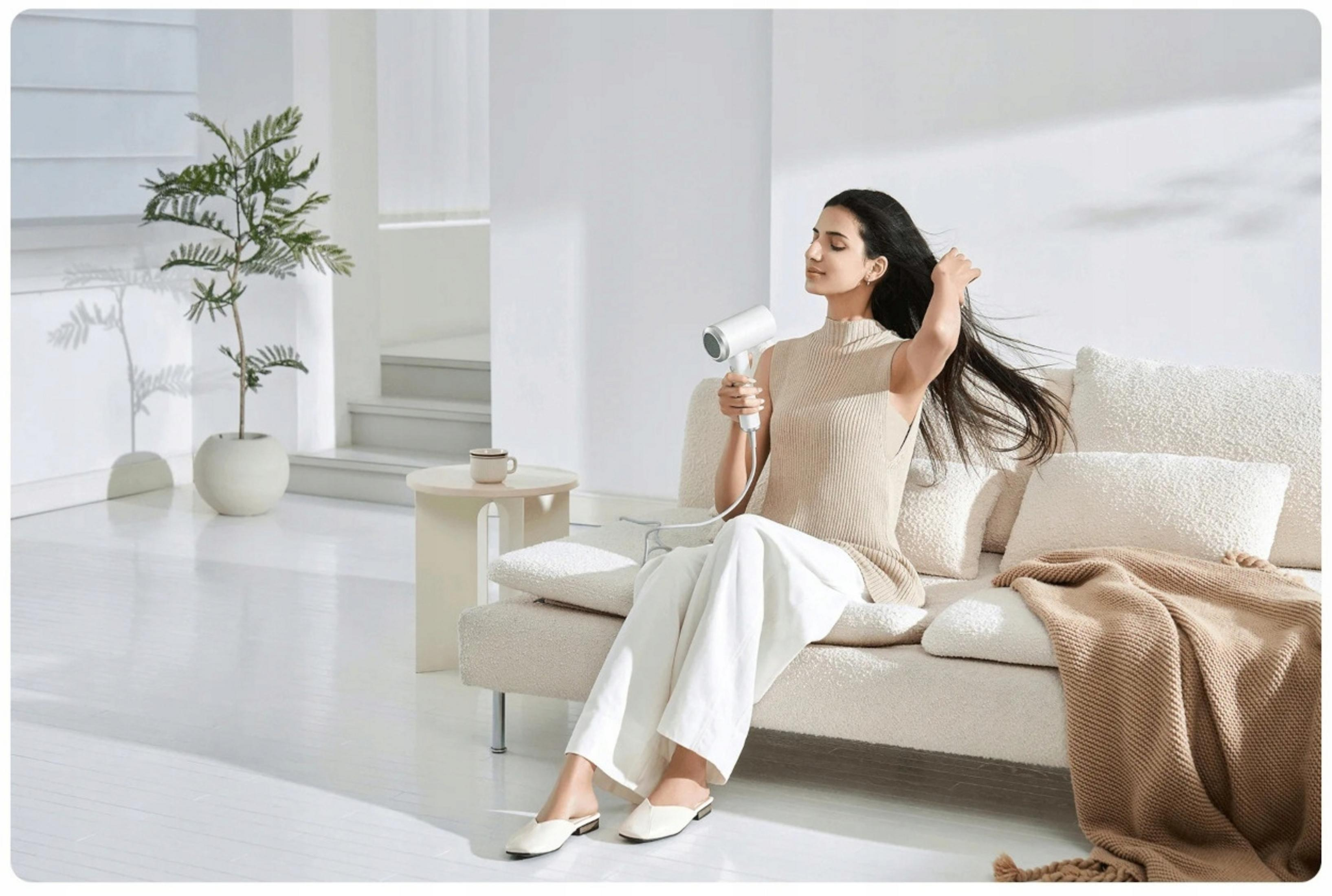 Kobieta z suszarką siedzi na kanapie. Suszy włosy. Pokój jest biały.