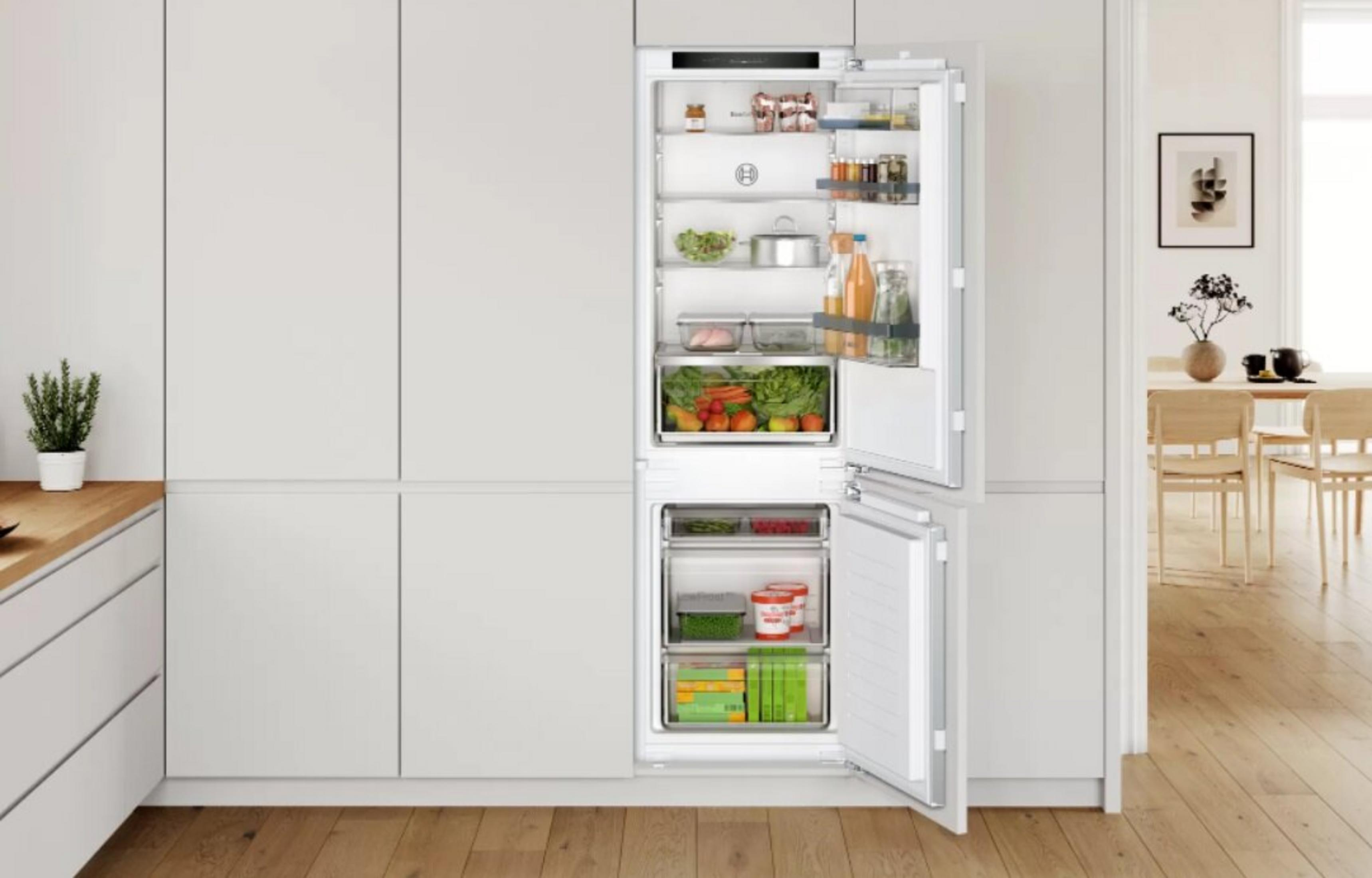 Otwarta biała lodówka wypełniona jedzeniem. Kuchnia w tle, drewniana podłoga i mała roślina na blacie.