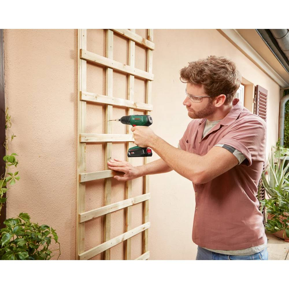 Ein Mann bohrt ein Holzgitter an eine rosa Wand. Er trägt eine Schutzbrille und ein rosa Hemd. Ein grüner Bohrer wird verwendet.