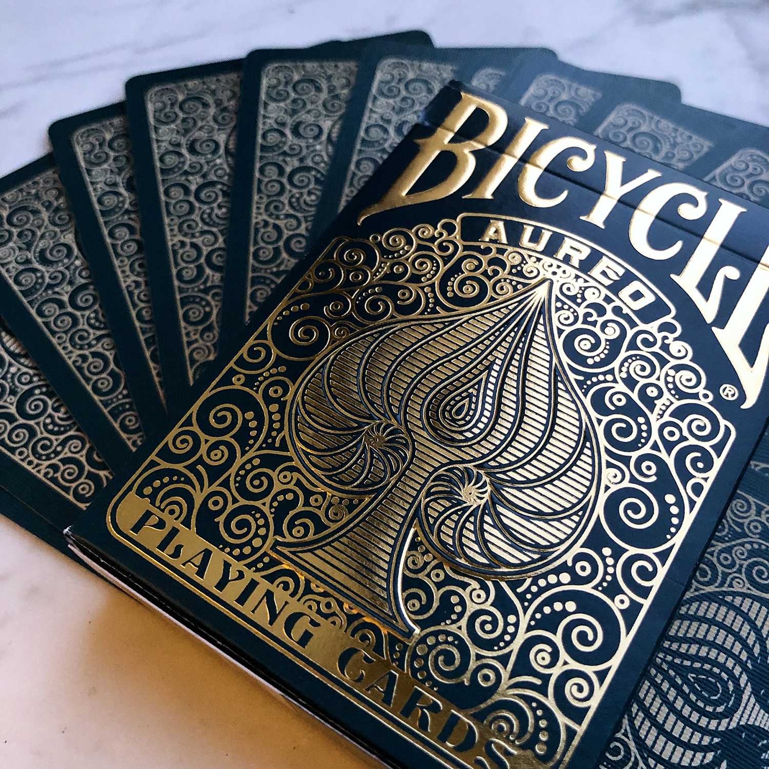Ein Satz Bicycle Aureo Spielkarten ist aufgefächert. Die Karten sind dunkelblau mit goldenen Akzenten, und die Schachtel ist sichtbar.