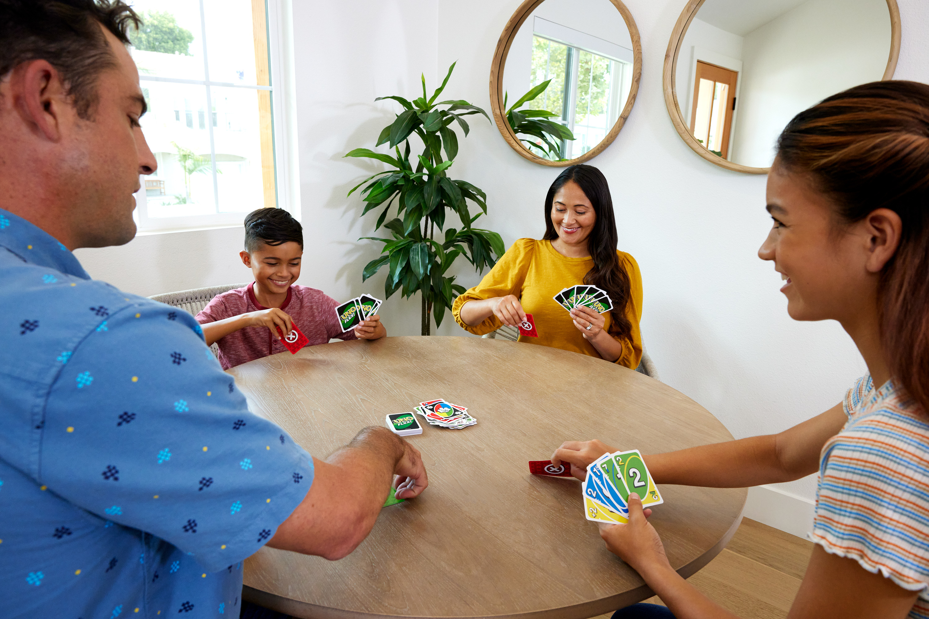 Quatre personnes jouent au Uno à une table ronde. Les cartes sont dans leurs mains. Une plante et des miroirs sont en arrière-plan.