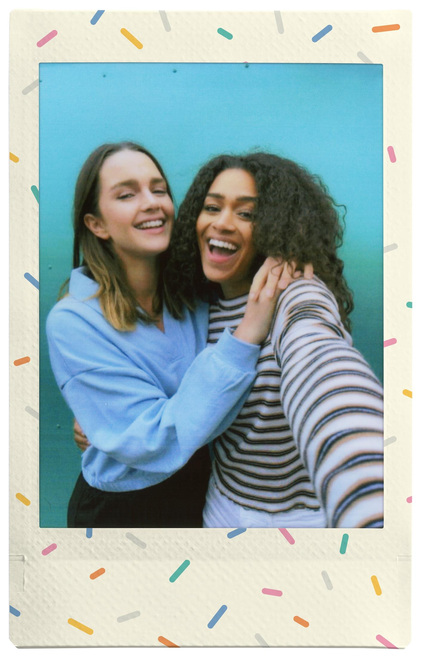 Dwie uśmiechnięte kobiety obejmują się, jedna w niebieskim, druga w paski, na tle turkusowego tła. Obraz jest w ramce polaroid.