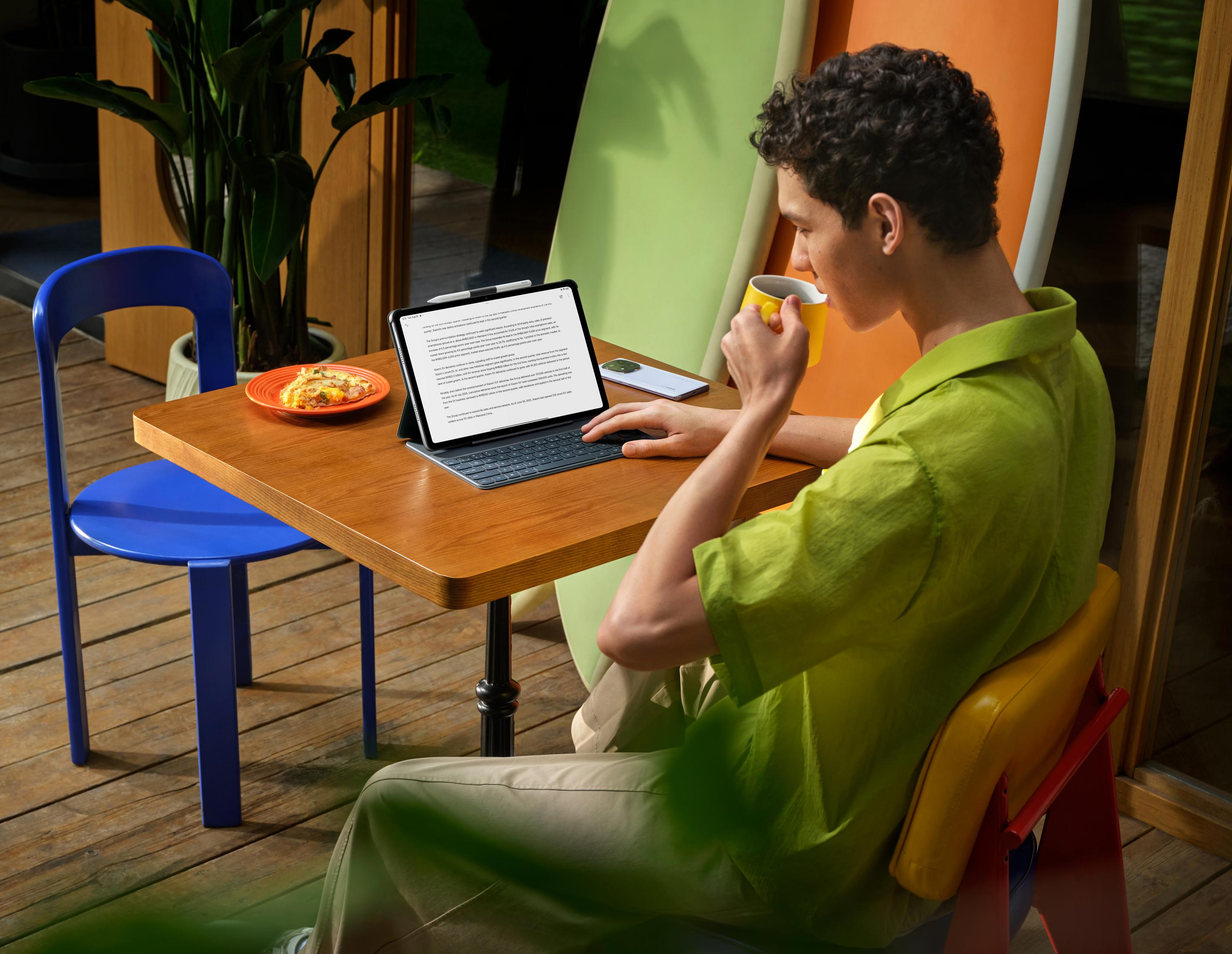 Mann benutzt Tablet, trinkt aus gelbem Becher. Pizza auf Teller, blauer Stuhl, Pflanze, Holztisch.