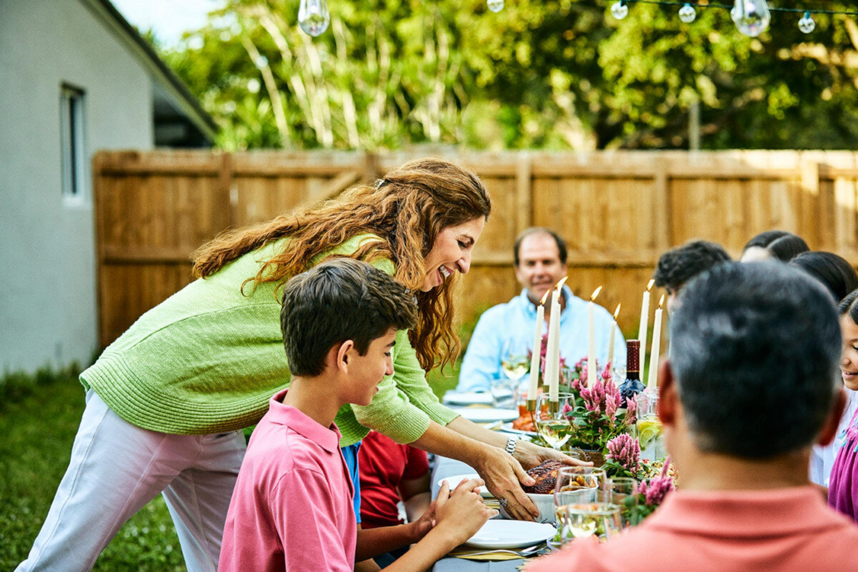Familie beim Abendessen im Freien. Frau serviert Essen. Tisch mit Kerzen, Blumen und Getränken. Menschen im Hintergrund.