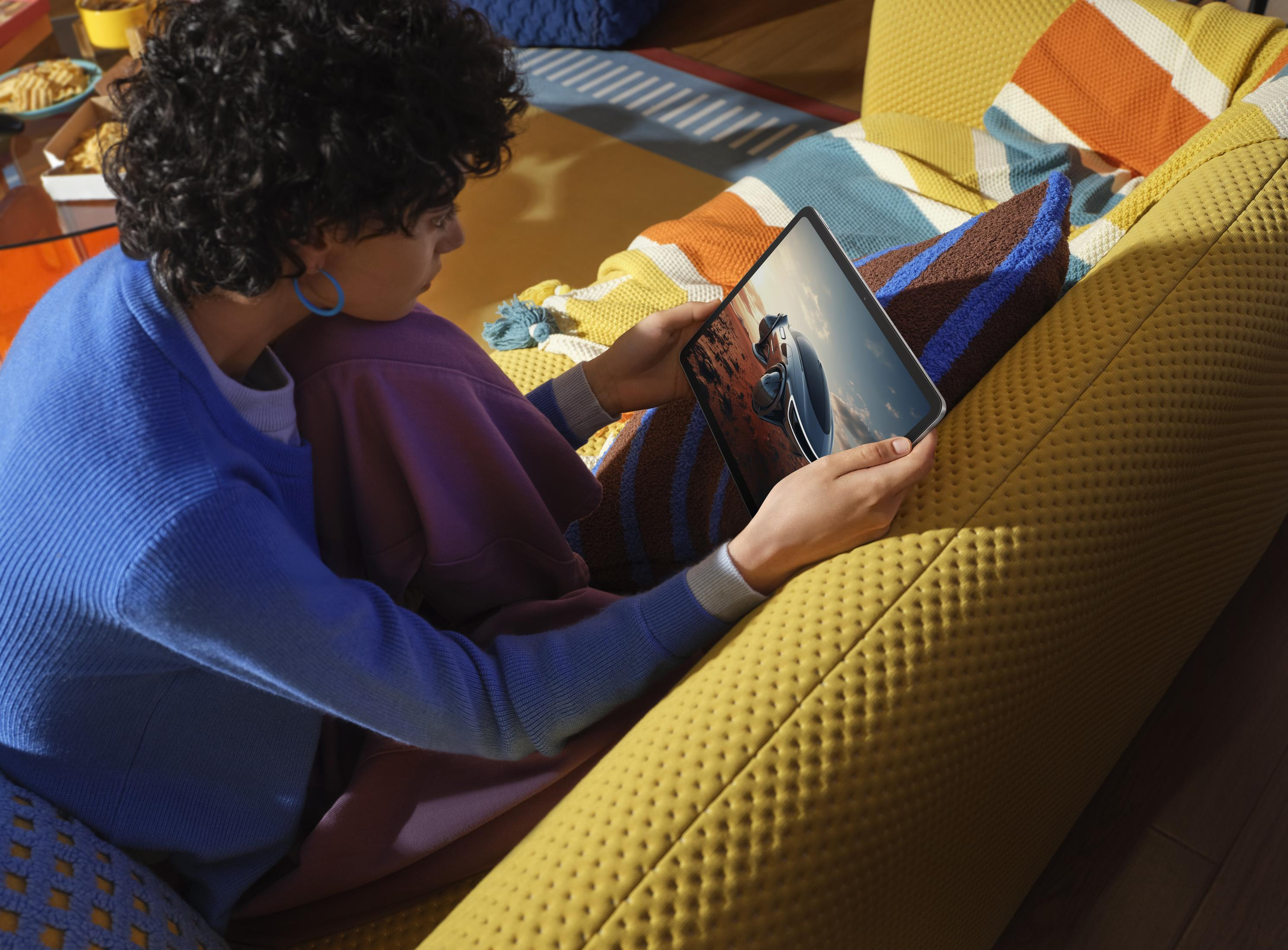 Osoba z kręconymi włosami patrzy na tablet. Siedzi na żółtej sofie z kolorowymi poduszkami.