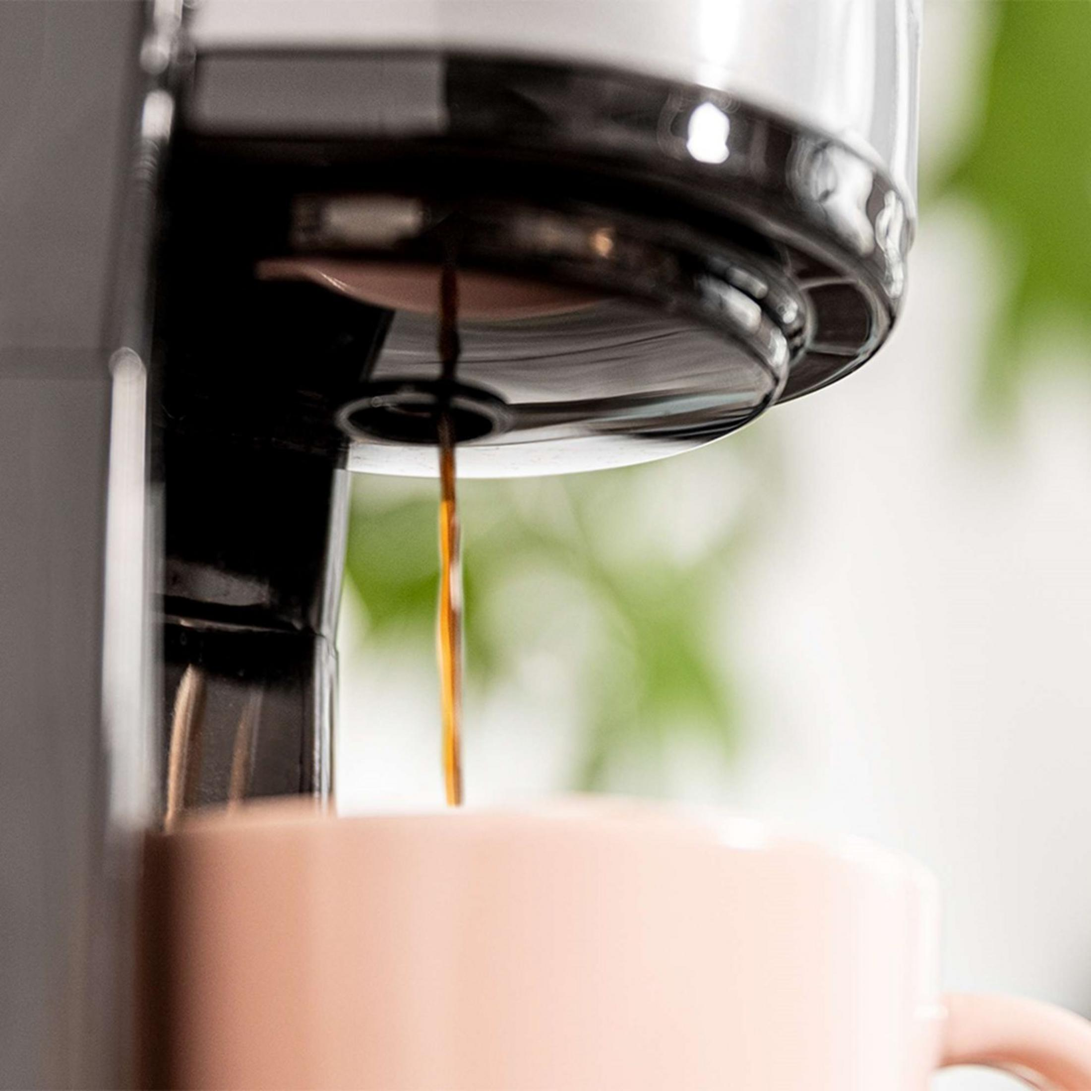 Kawa zaparzająca się do różowego kubka. Ekspres do kawy w tle. Zielone tło.