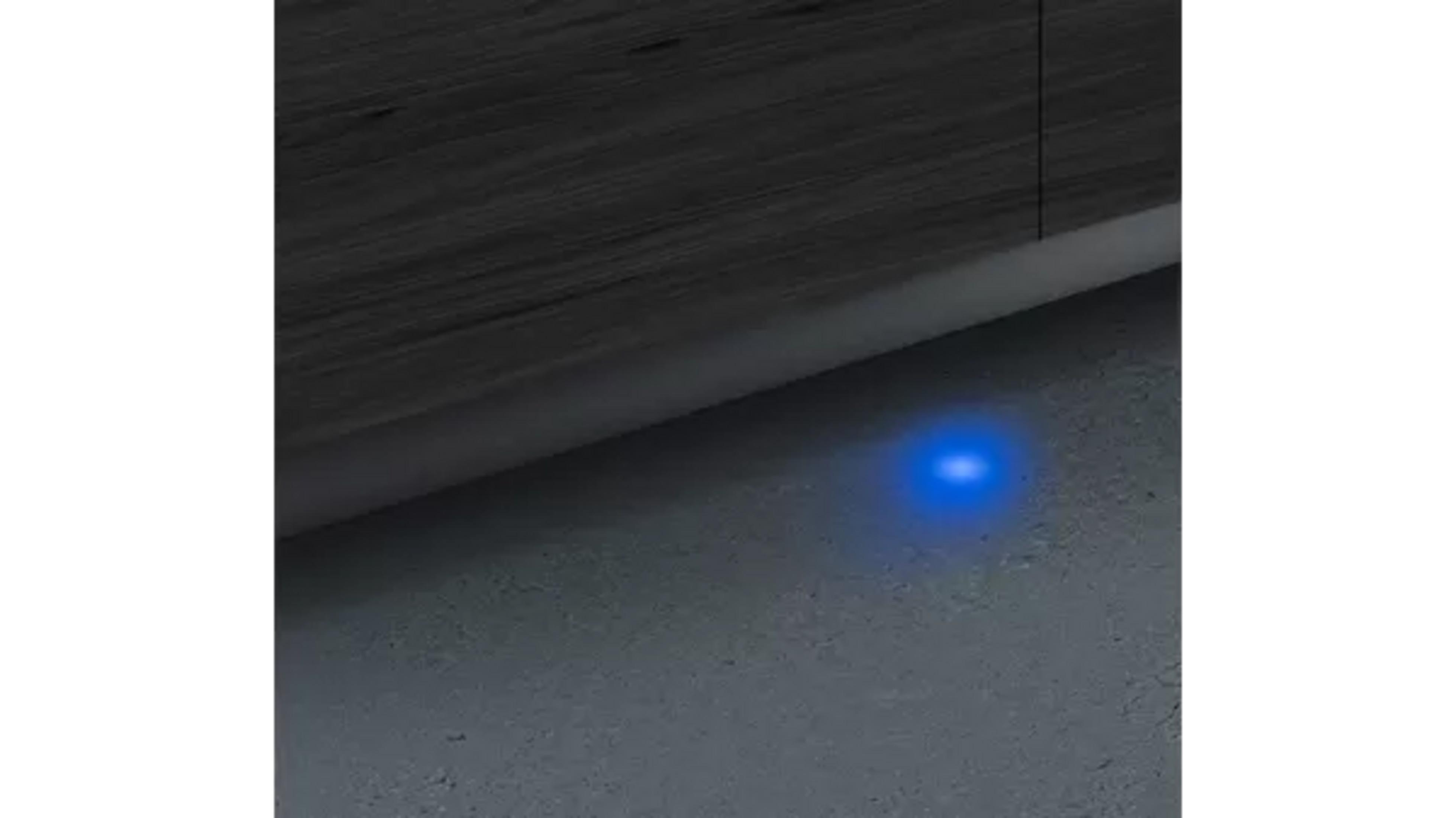 Une surface en bois sombre et un sol gris sont visibles. Une petite lumière bleue illumine le sol, près du bord.