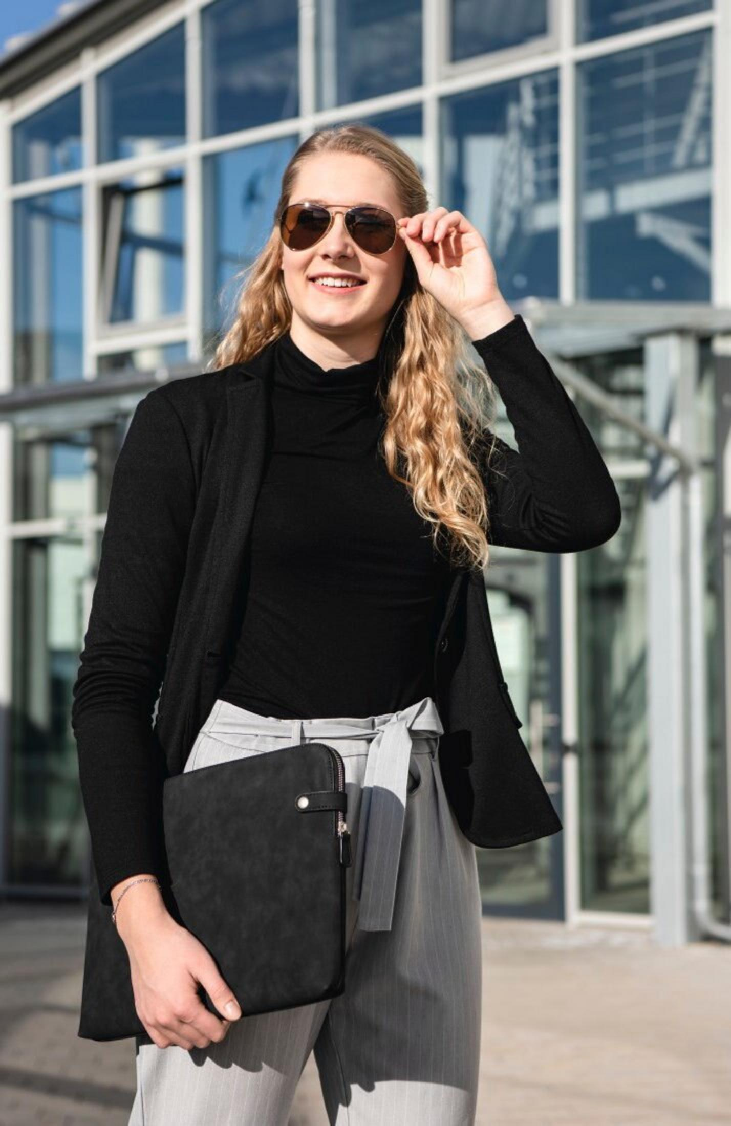 Kobieta w okularach przeciwsłonecznych i czarnym stroju, trzymająca czarną teczkę, uśmiechnięta przed szklanym budynkiem.