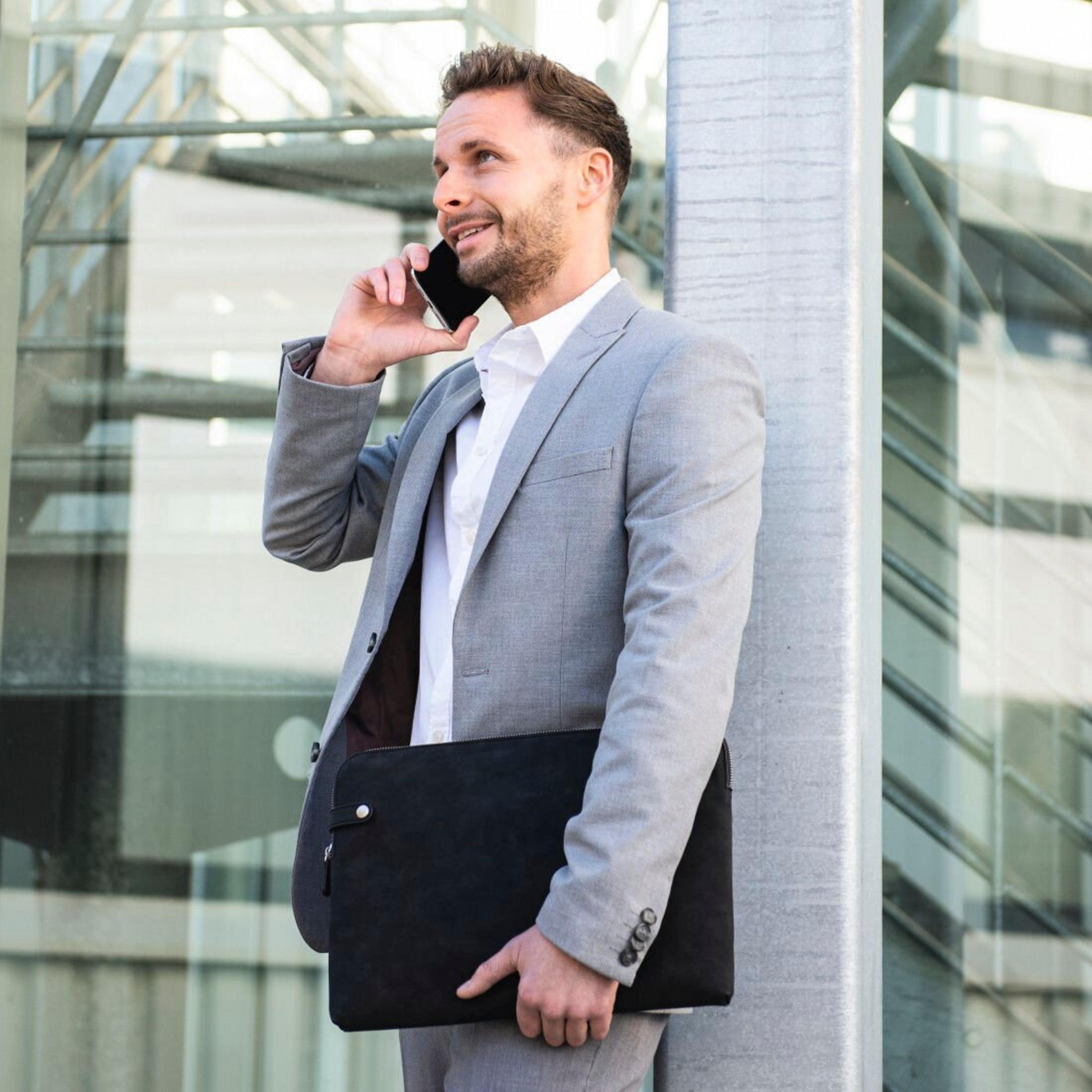 Mężczyzna w szarym garniturze rozmawia przez telefon, trzymając czarną teczkę. Opiera się o słup obok szklanego budynku.