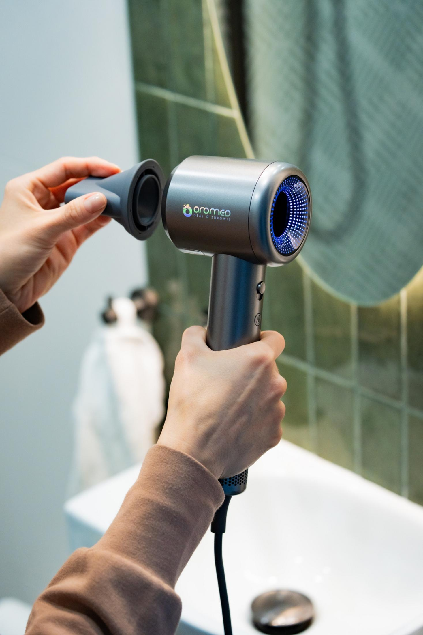 Osoba trzyma srebrną suszarkę do włosów. Jedna ręka trzyma nasadkę dyszy, druga uchwyt. Tło łazienki.