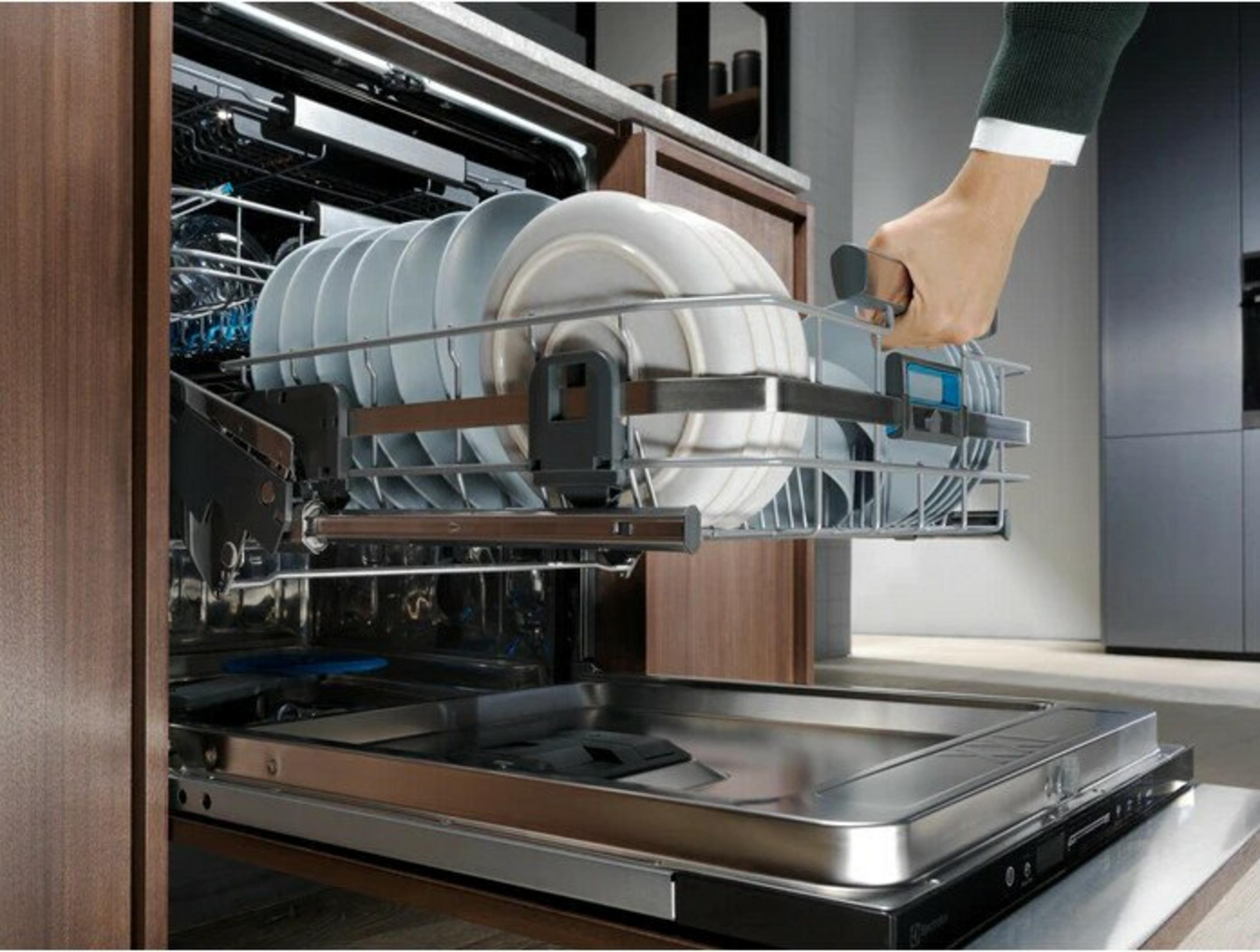 Zmywarka jest otwarta, załadowana talerzami. Ręka wyciąga ruszt. Otoczenie kuchni.