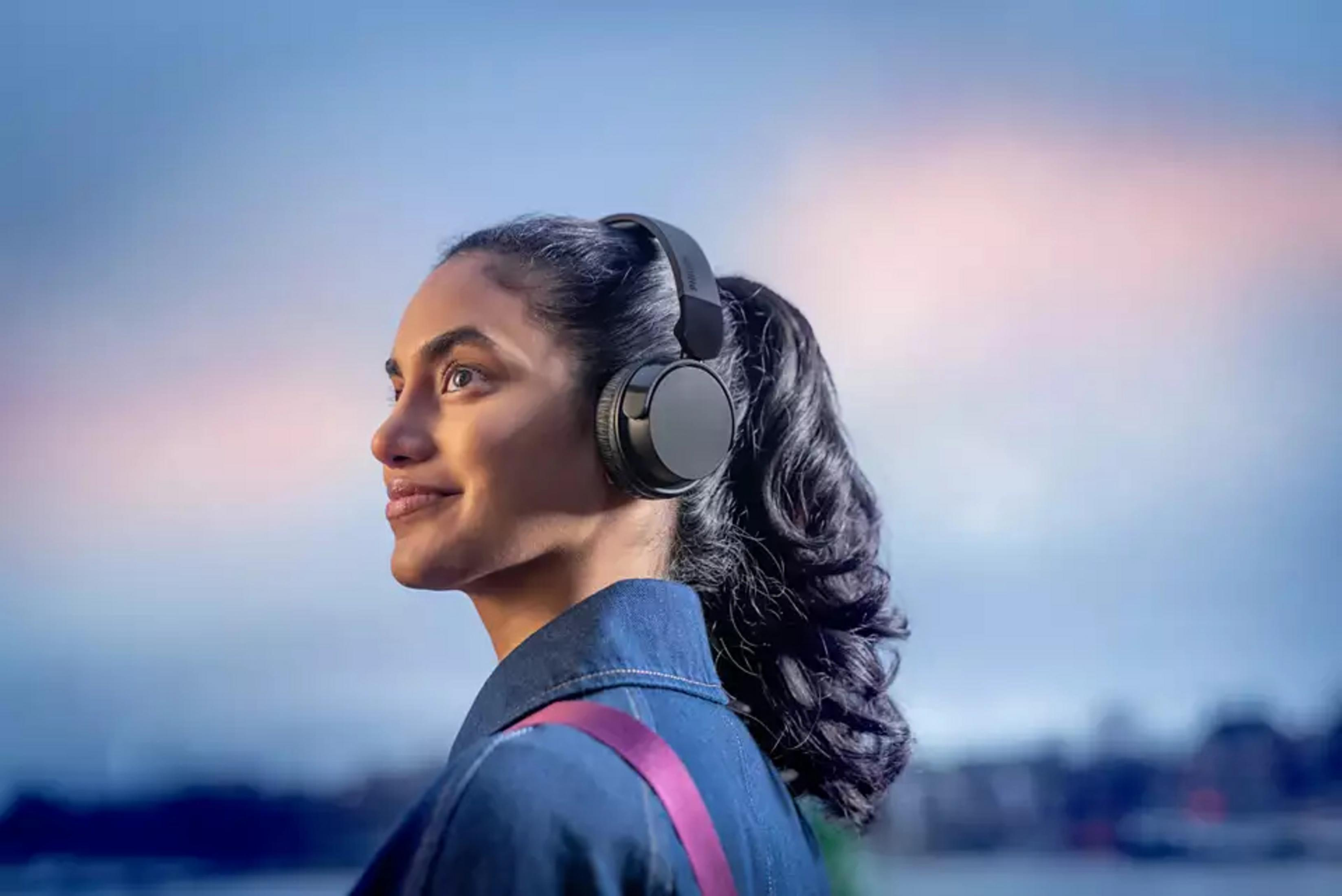 Mujer con auriculares, chaqueta vaquera azul y una correa rosa. Cielo al fondo.
