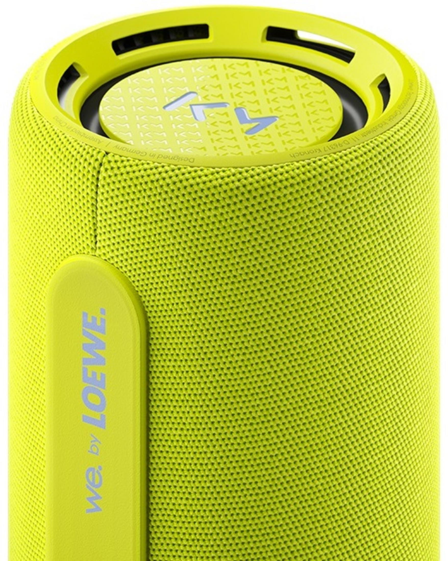 Ein zylindrischer Lautsprecher in Limettengrün mit dem Markennamen 'We. by Loewe'. Die Oberseite hat ein Logo und Text. Der Lautsprecher ist strukturiert.