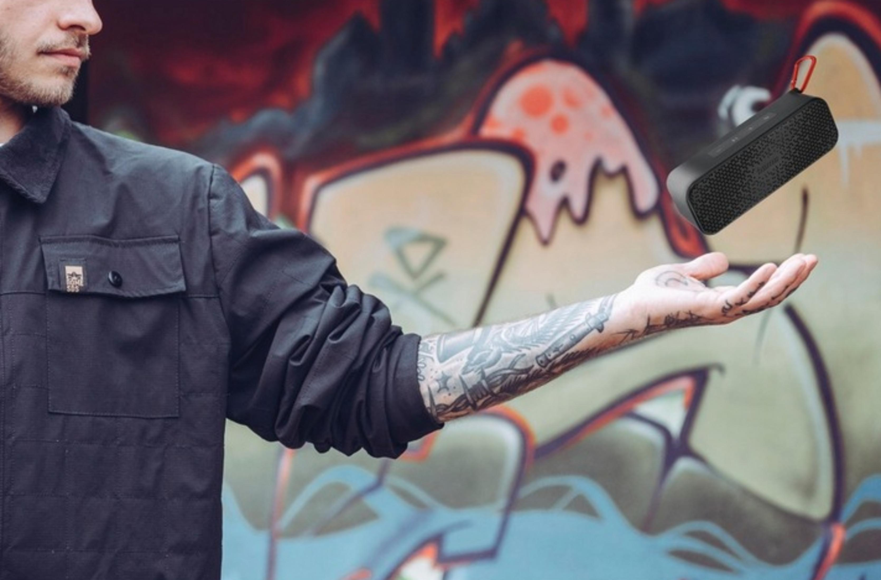 Mężczyzna z tatuażami trzymający czarny głośnik z czerwoną pętlą. Tło graffiti.