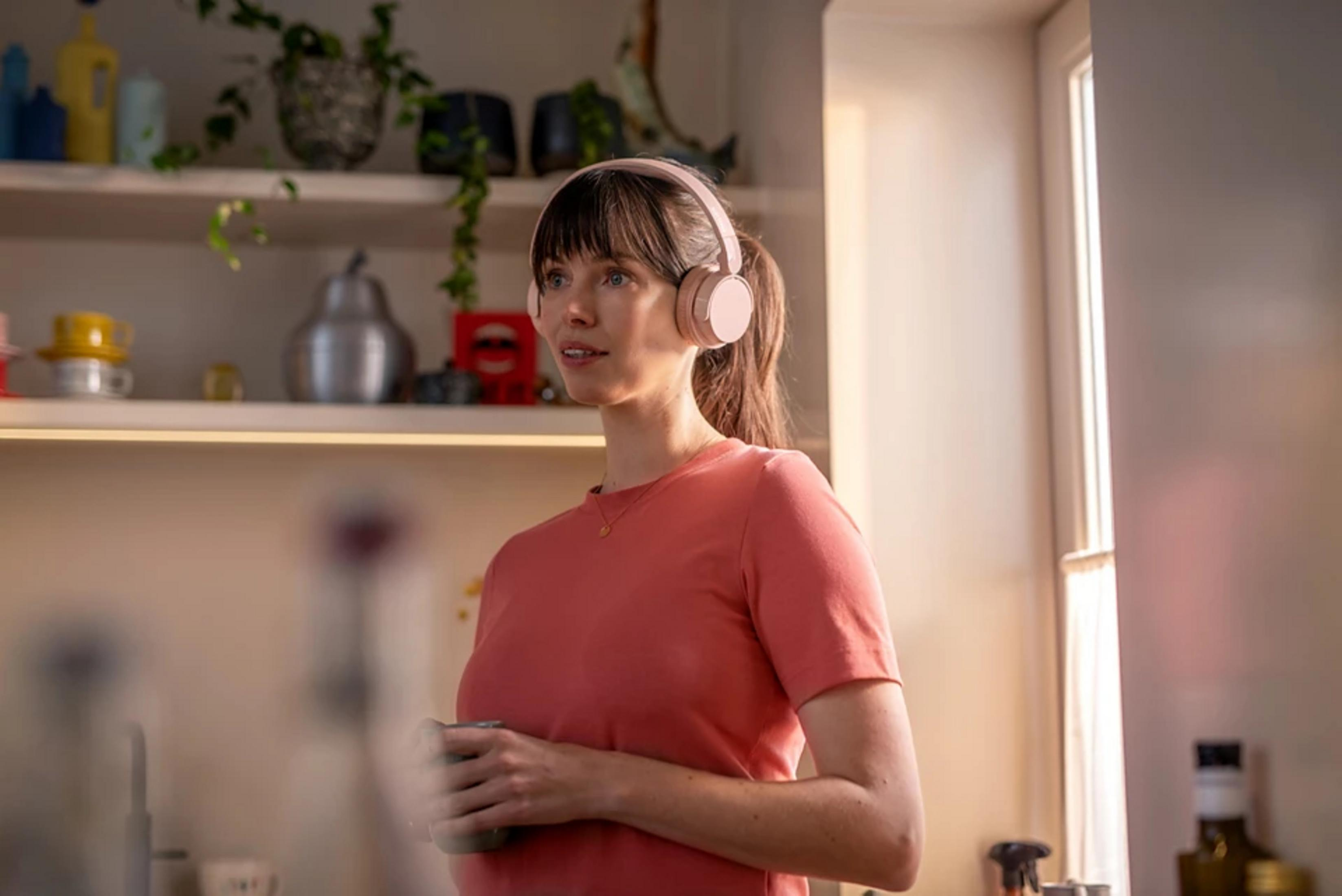 A woman wearing pink headphones and shirt holds a mug. Shelves hold plants and containers. A window is in the background.