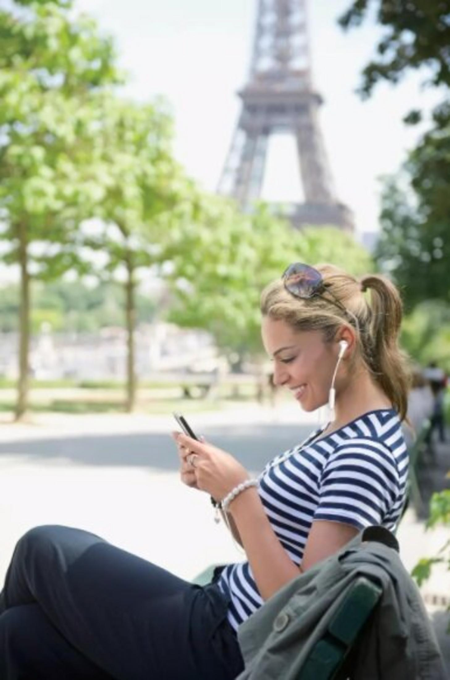 Femme en chemise rayée écoute de la musique, utilise un téléphone, sourit. Tour Eiffel en arrière-plan. Lunettes de soleil sur les cheveux.