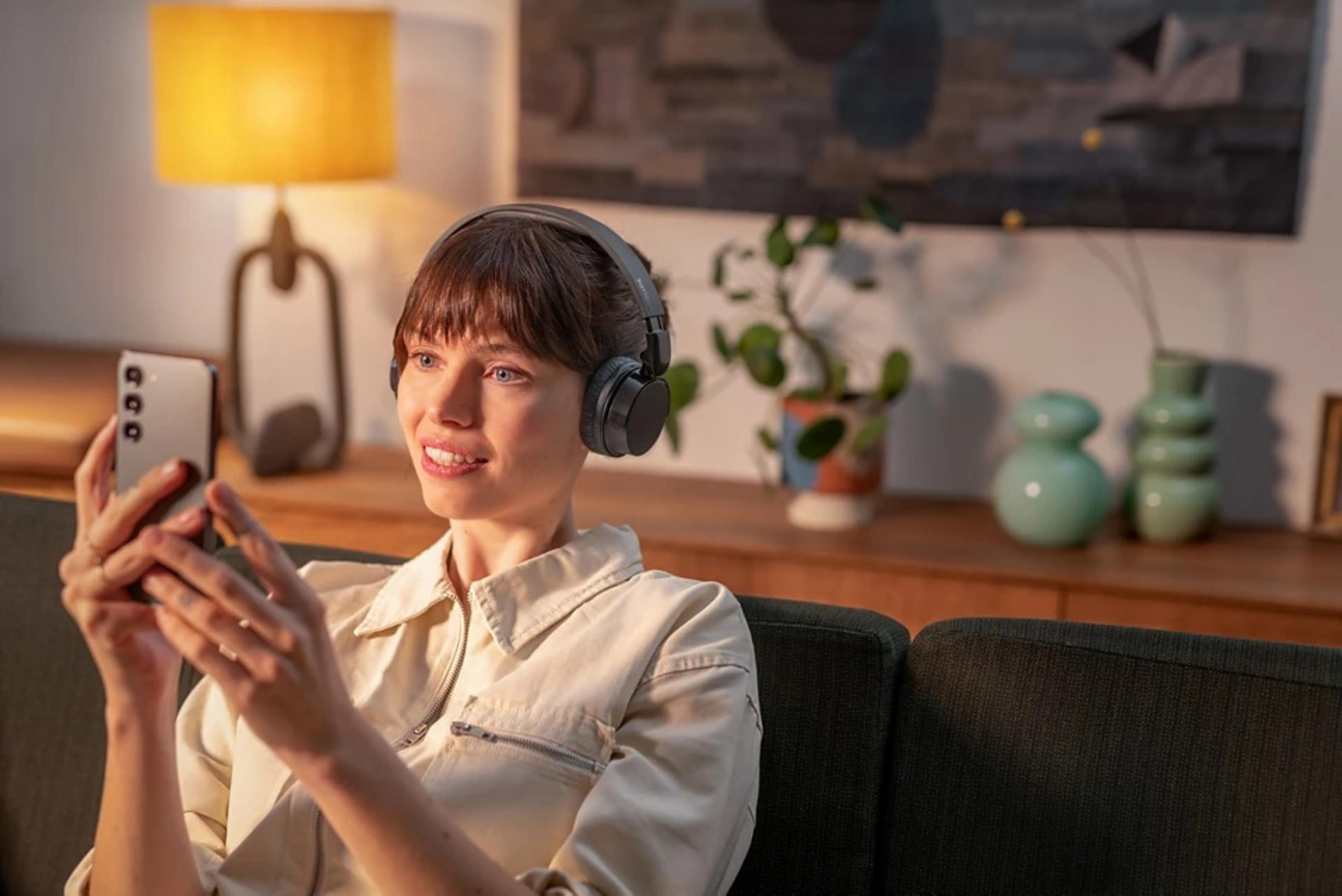 Eine Frau mit Kopfhörern schaut auf ihr Handy. Sie sitzt auf einem Sofa. Im Hintergrund eine Lampe und Deko-Artikel.