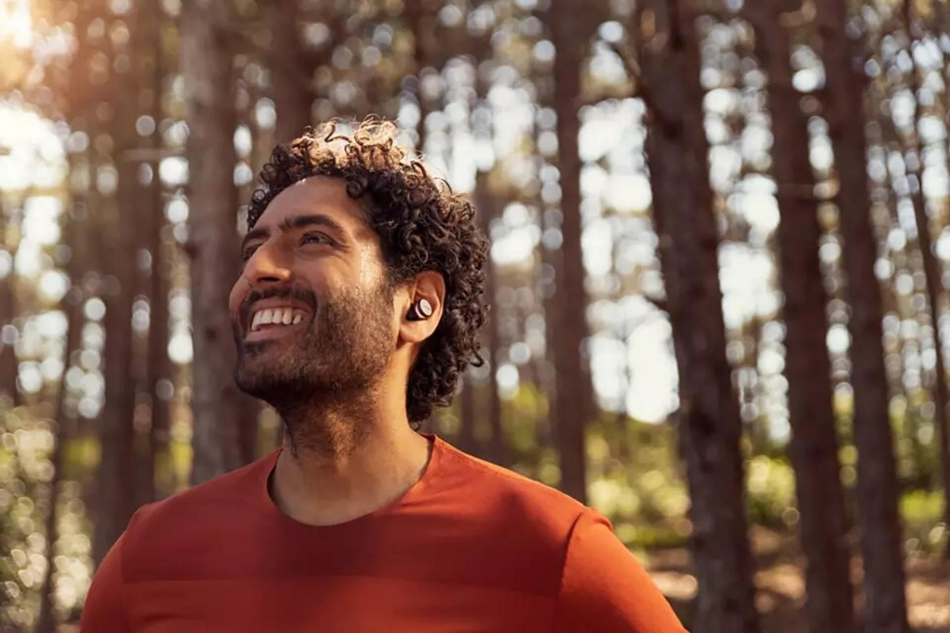 Mężczyzna uśmiecha się, nosząc słuchawki i pomarańczową koszulę, w lesie.