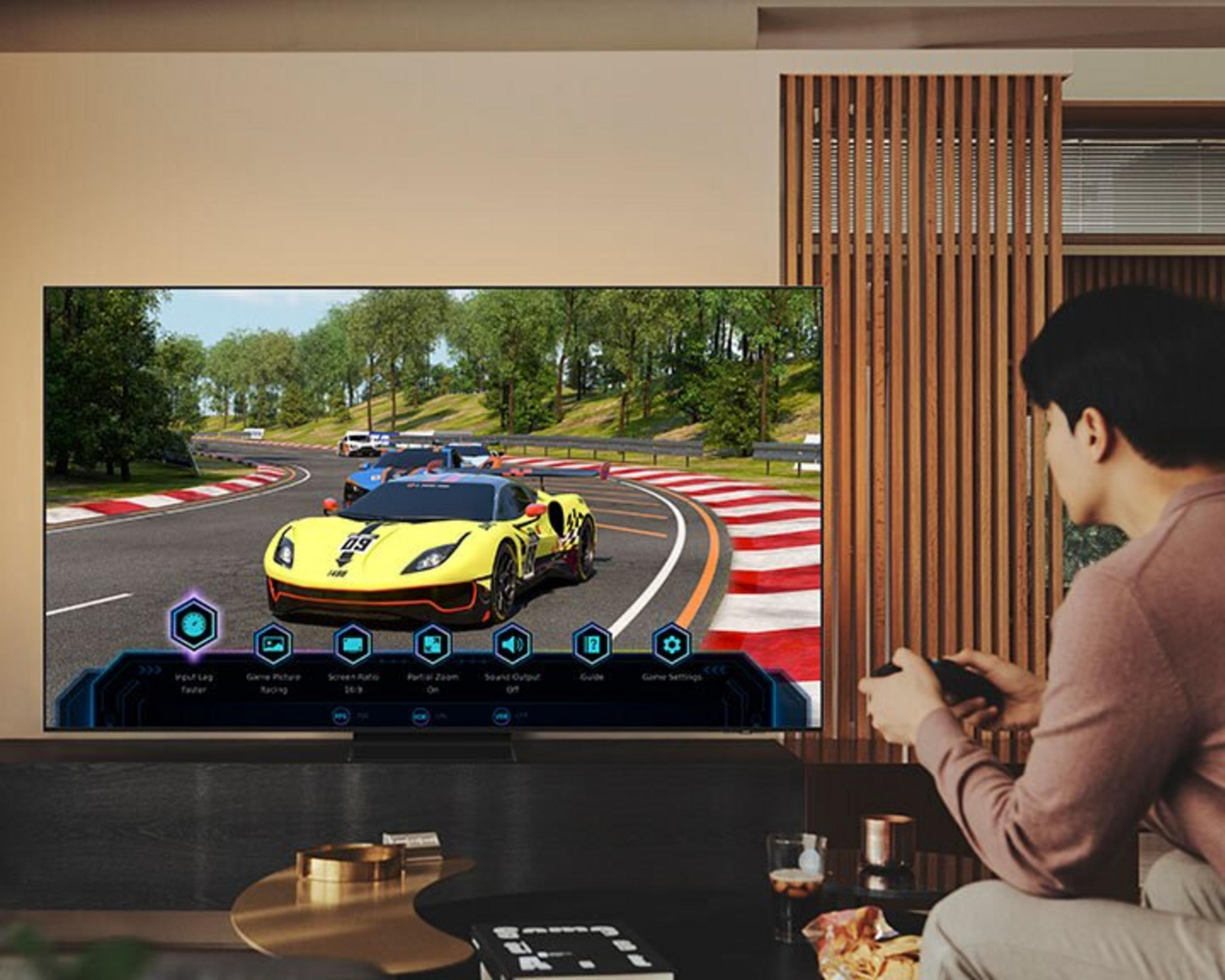 Una persona juega un juego de carreras de coches en un televisor grande. Un coche amarillo lidera la carrera. La persona sostiene un mando.