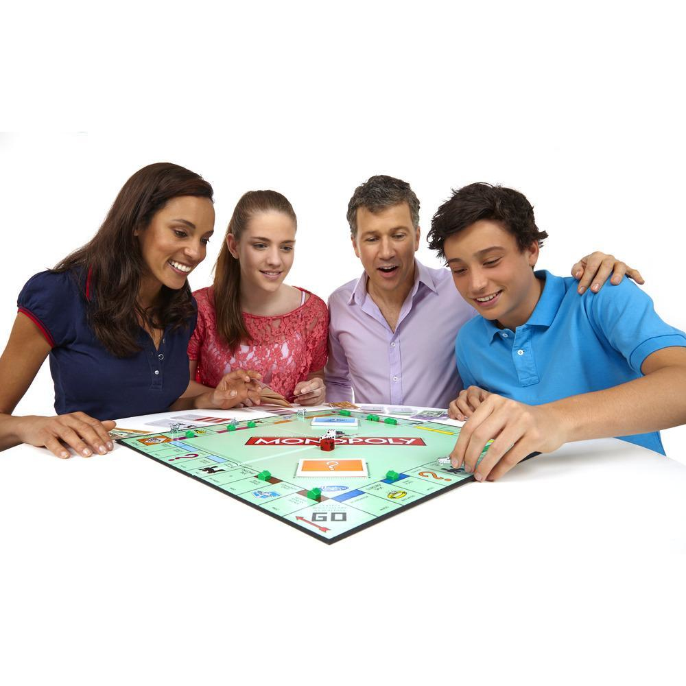 Vier Personen spielen Monopoly. Sie stehen lächelnd um das Spielbrett herum. Das Brett ist grün mit rotem und weißem Text.