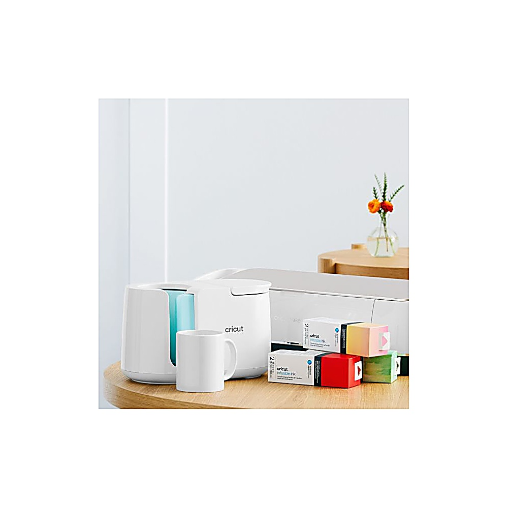 Eine weiße Cricut-Maschine, eine Tasse und Kartons stehen auf einem Holztisch. Eine Vase mit Blumen steht im Hintergrund.