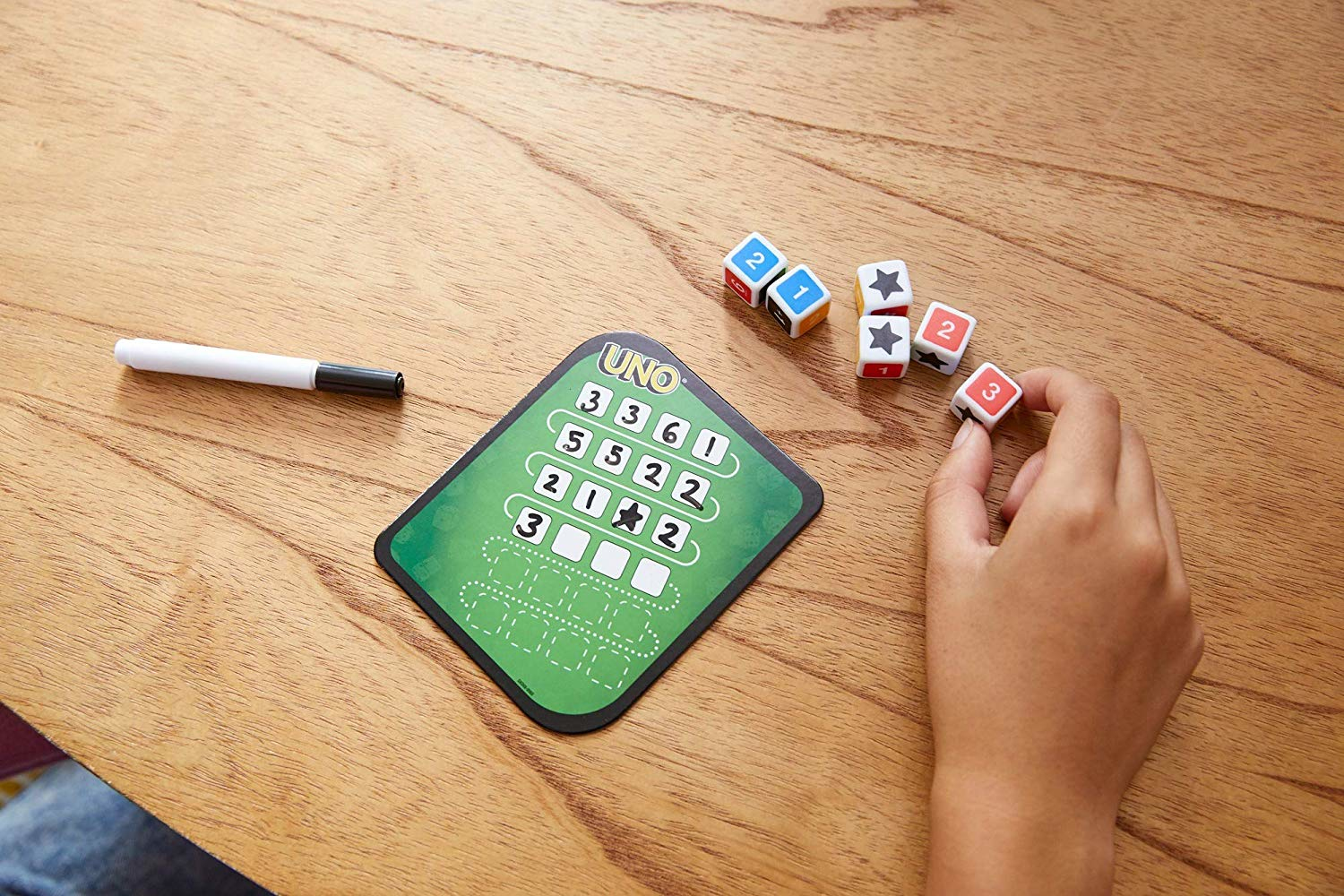 UNO-Spiel auf einem Holztisch. Enthält Würfel, einen Marker und eine UNO-Karte. Hand hält einen Würfel.