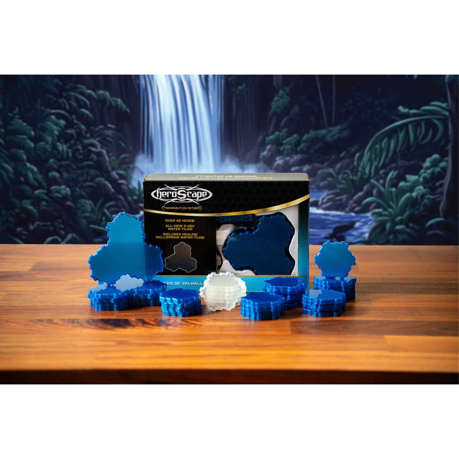 Eine Heroscape-Box mit blauen und weißen sechseckigen Fliesen auf einem Holztisch. Ein Wasserfall und grünes Laub sind im Hintergrund.