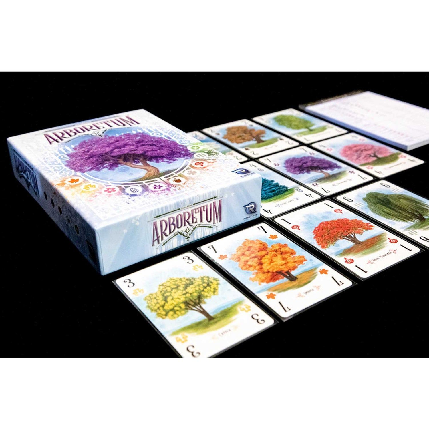 Ein Brettspiel, Box und Karten liegen auf schwarzem Untergrund. Die Box zeigt ein lilafarbenen Baum. Karten zeigen verschiedene Bäume.