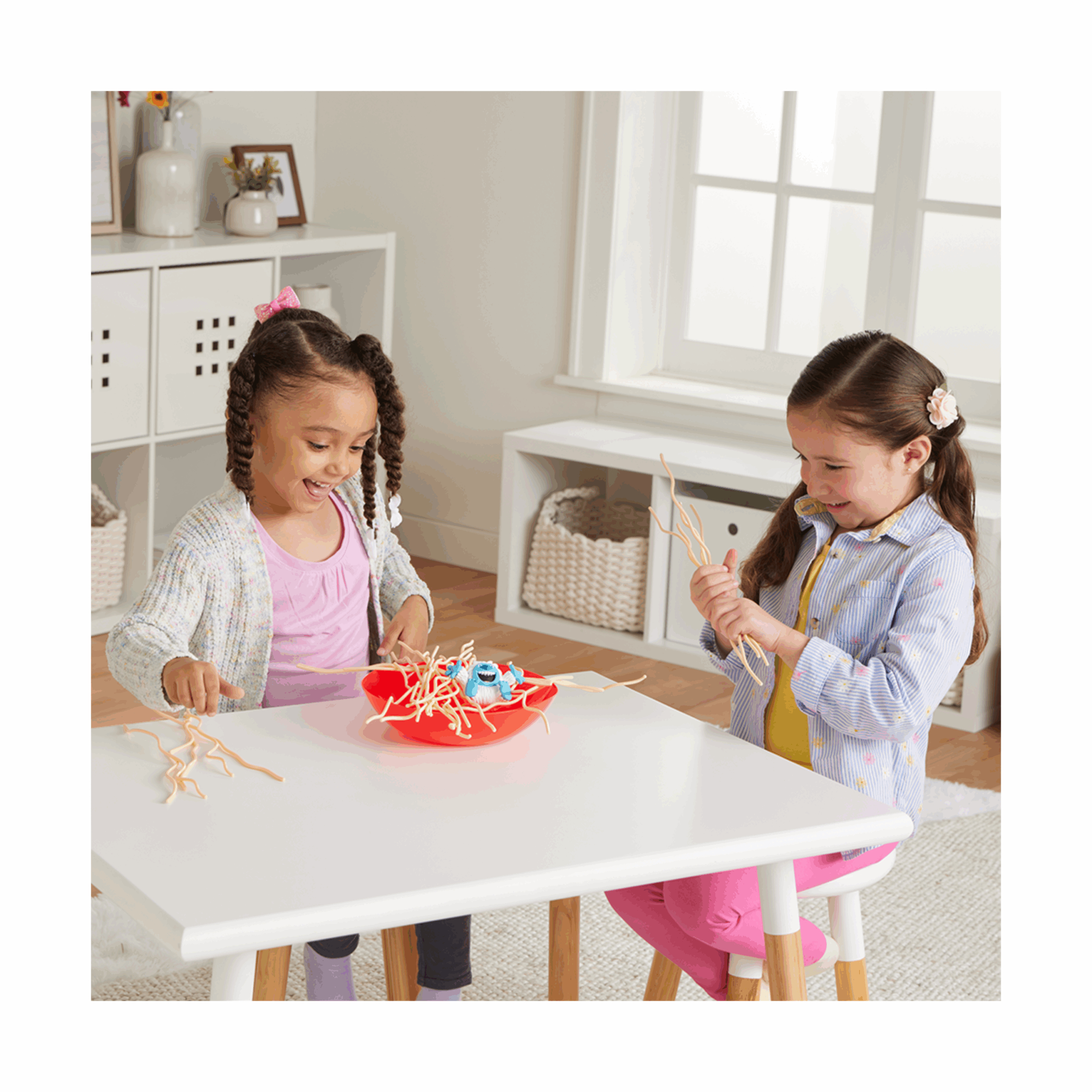 Deux filles jouant à un jeu. Un bol rouge avec des spaghettis et un yéti jouet sur une table blanche.