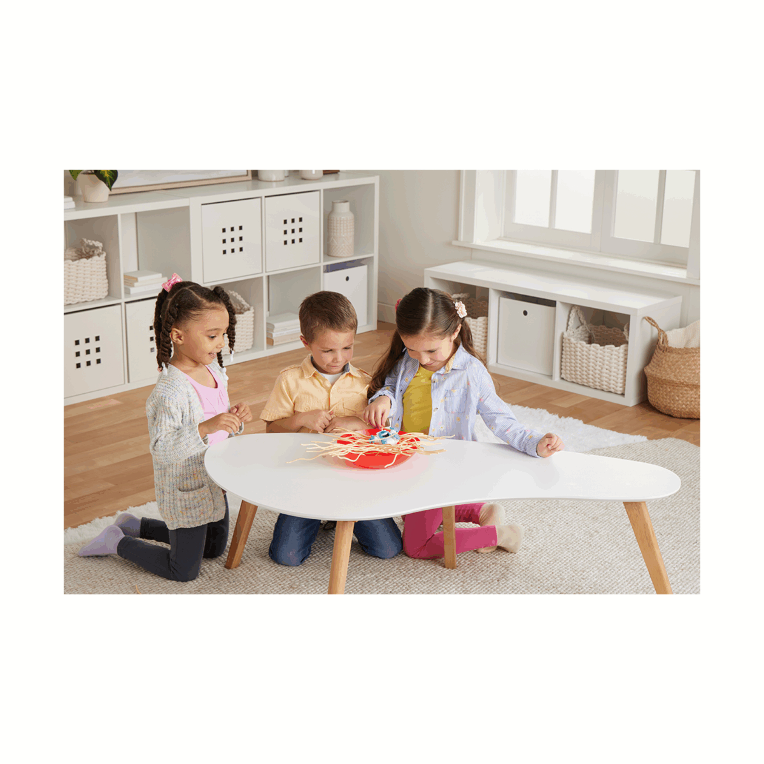 Trois enfants jouent à un jeu sur une table blanche. Un bol rouge avec des spaghettis est au centre.