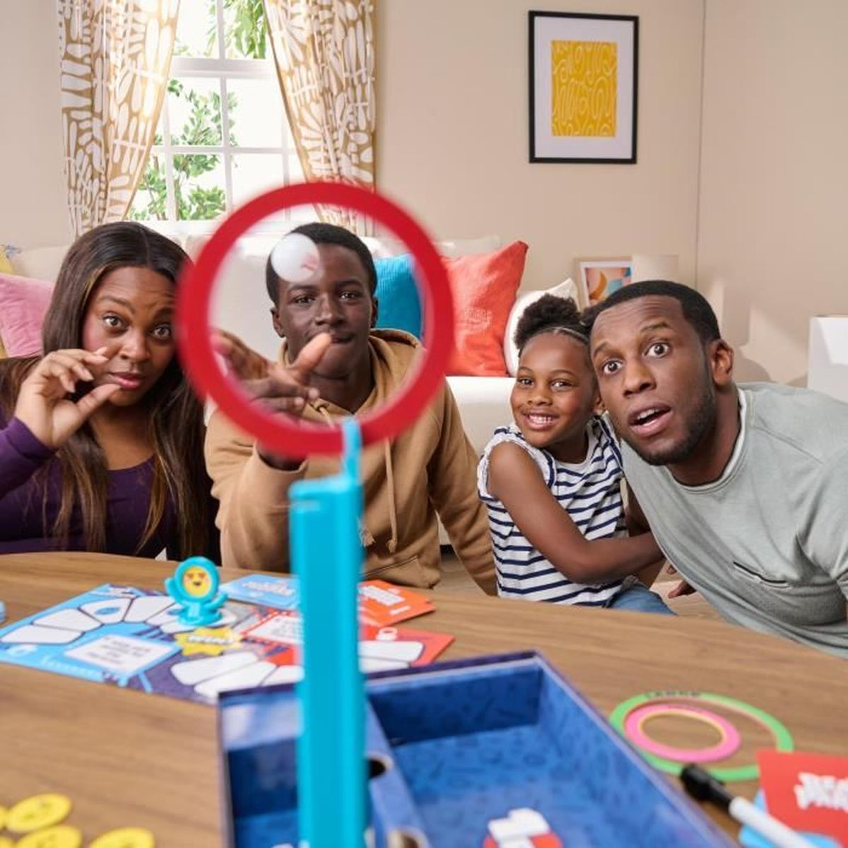 Een gezin speelt een bordspel. Een rode cirkel omlijst een persoon. Anderen hebben verraste uitdrukkingen. Spelstukken en een raam zijn zichtbaar.