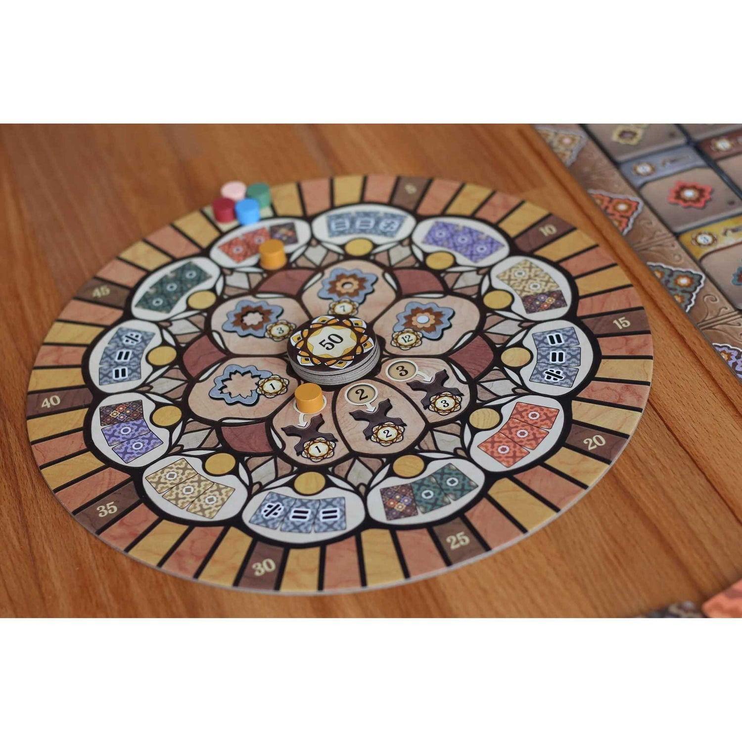 Ein kreisförmiges Brettspiel mit floralem Design liegt auf einem Holztisch. Es gibt Spielsteine ​​verschiedener Farben und eine Karte.