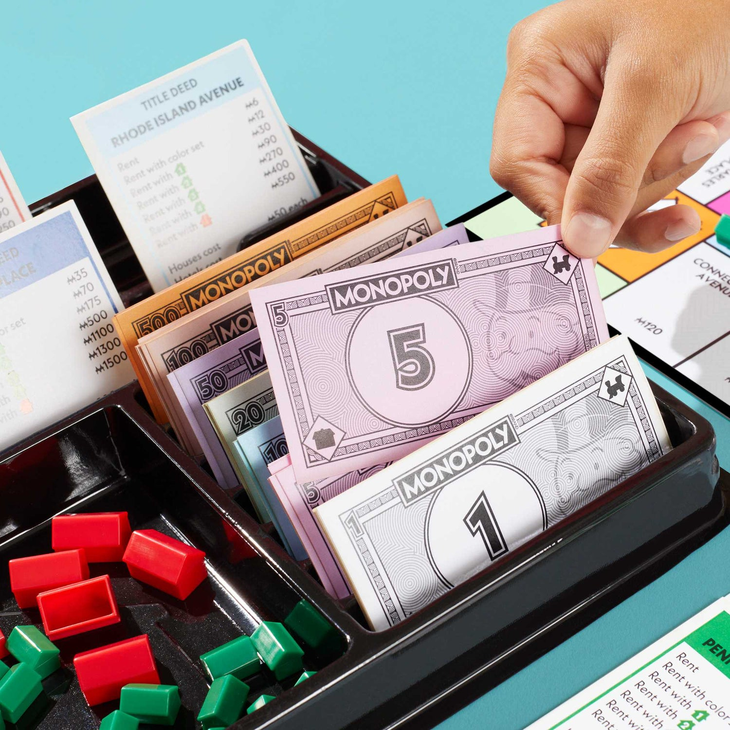 Una mano sosteniendo dinero de Monopoly sobre una bandeja con fichas y tarjetas de propiedad, sobre una superficie azul.