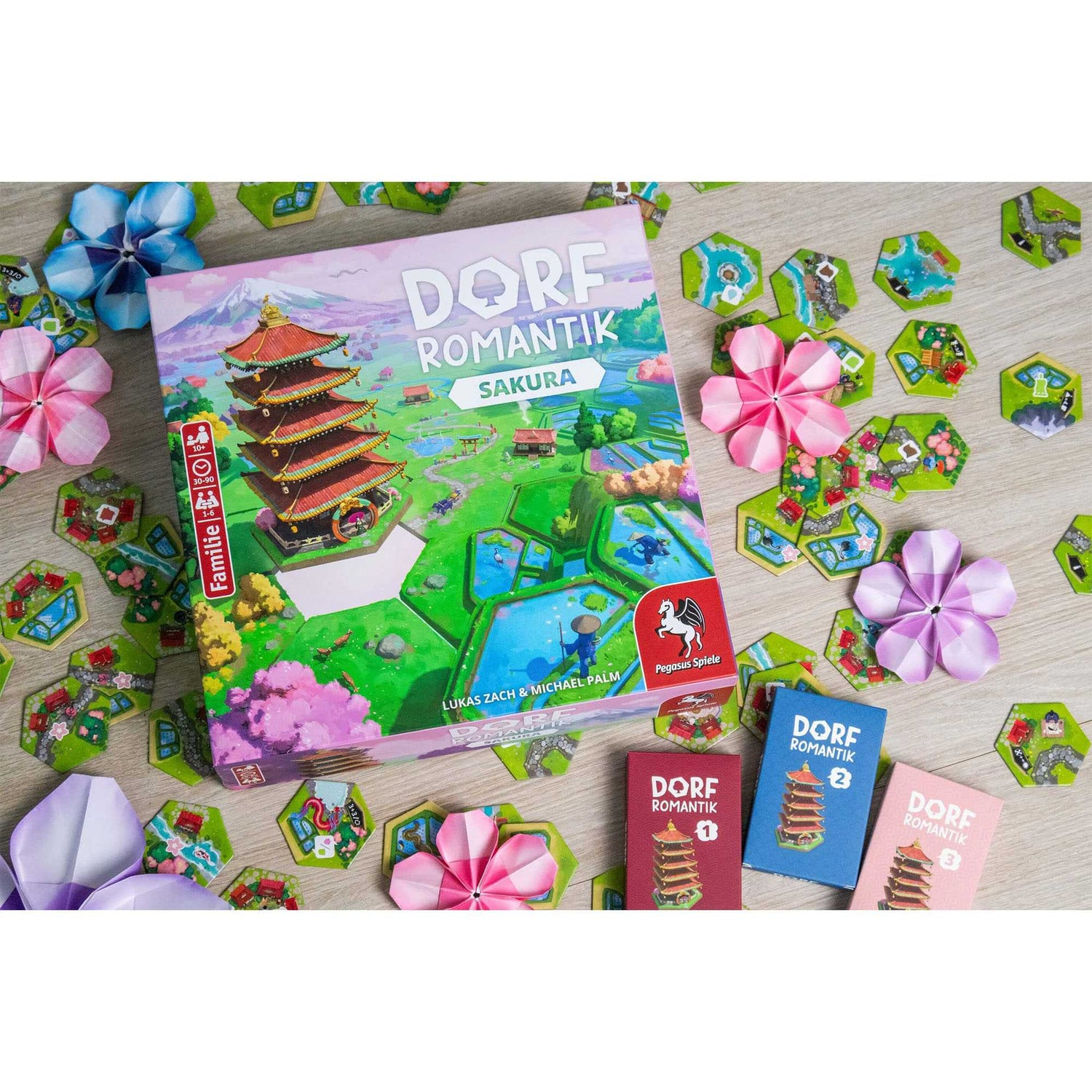 Ein Brettspiel namens 'Dorf Romantik Sakura' mit Spielsteinen und Origami-Blumen auf einer Holzoberfläche. Mehrere kleine Spielkarten sind vorhanden.